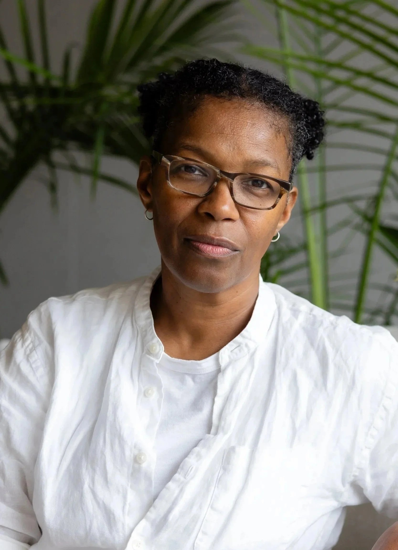 A woman with short curly hair, wearing glasses and a white button-up shirt, standing in front of a large green plant.