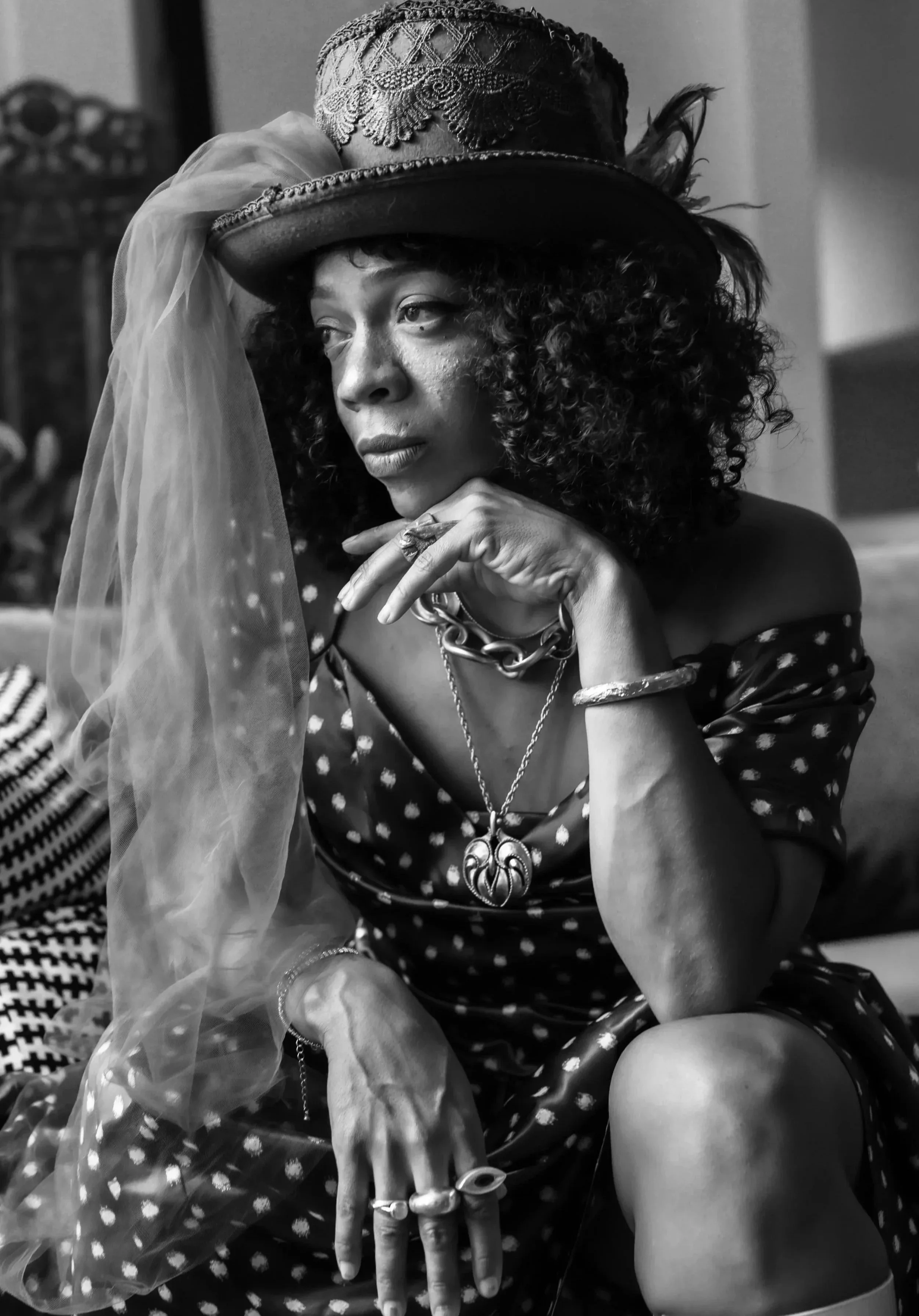 Black and white photo of a woman wearing a vintage hat with feathers and lace, a wig with tight curls, multiple necklaces, rings, and a patterned dress. She is seated with her hand resting on her face, gazing pensively.