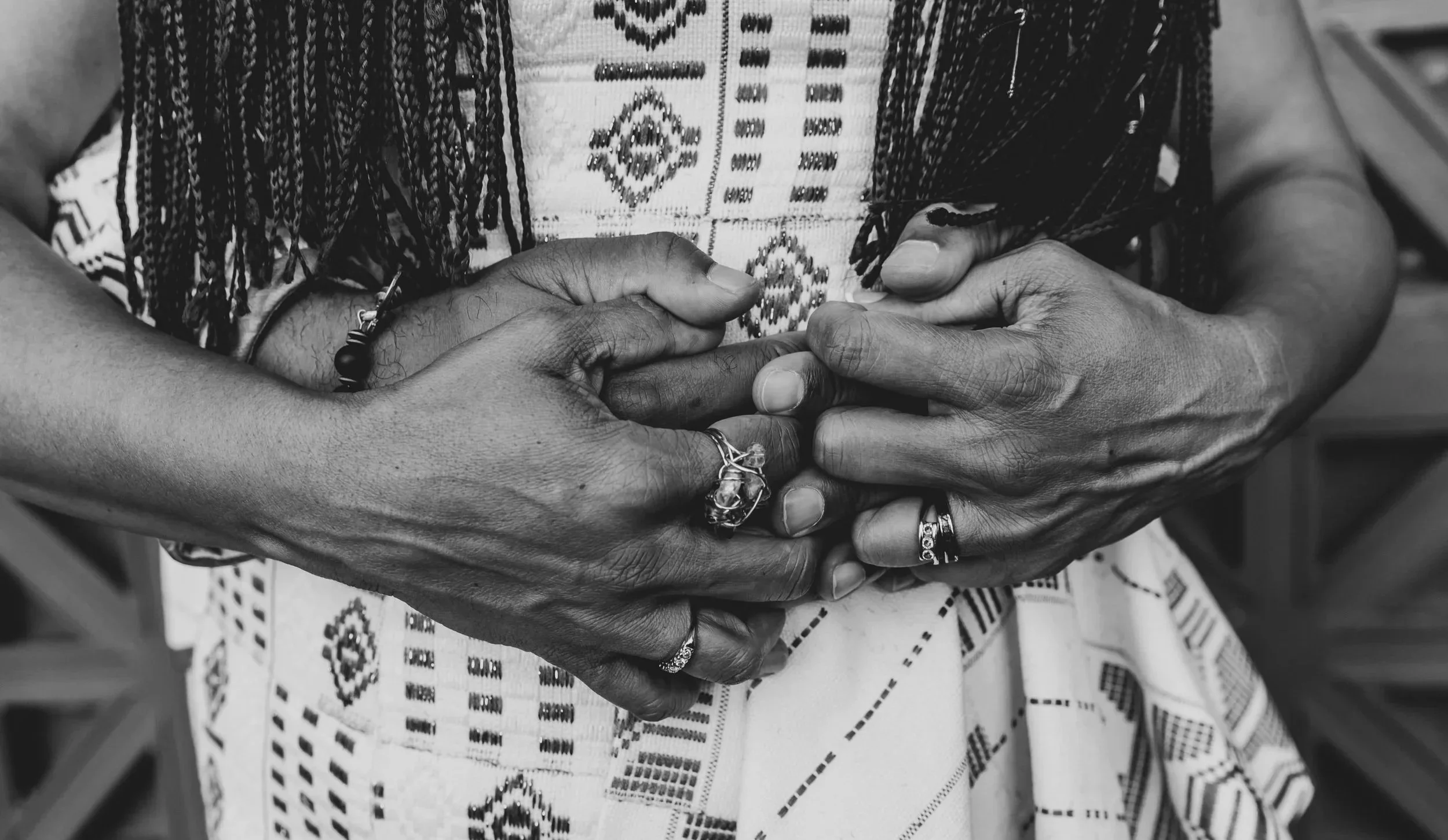 A person with dark skin wearing multiple rings on their fingers, holding or clasping their hands together, with a patterned fabric or clothing visible in the background.