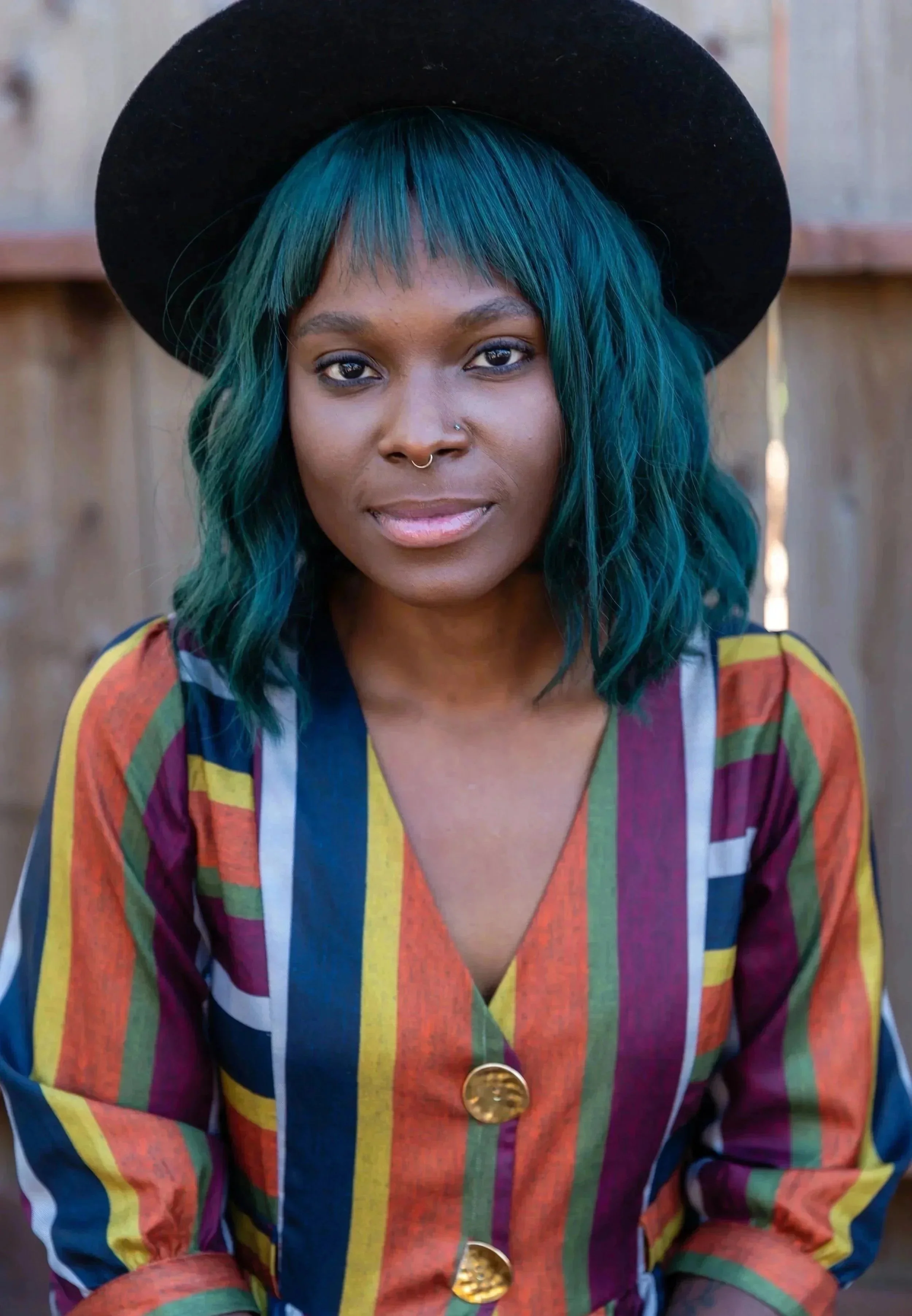 A woman with teal and blue striped hair, wearing a black wide-brimmed hat and a colorful, striped button-up shirt with gold buttons, in front of a wooden background.