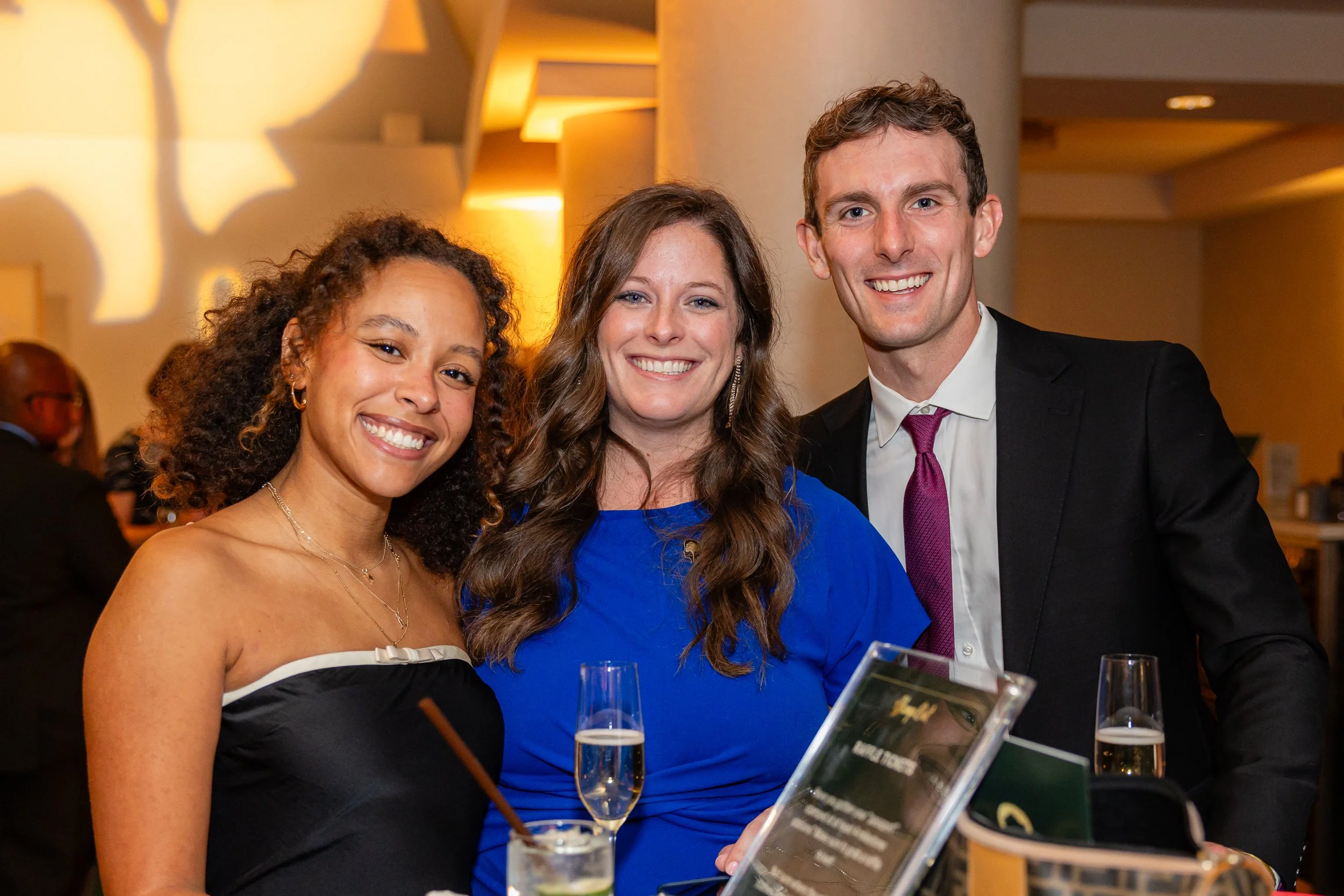 Three people smiling at a social event, standing close together, holding drinks, with a warm, softly lit background.