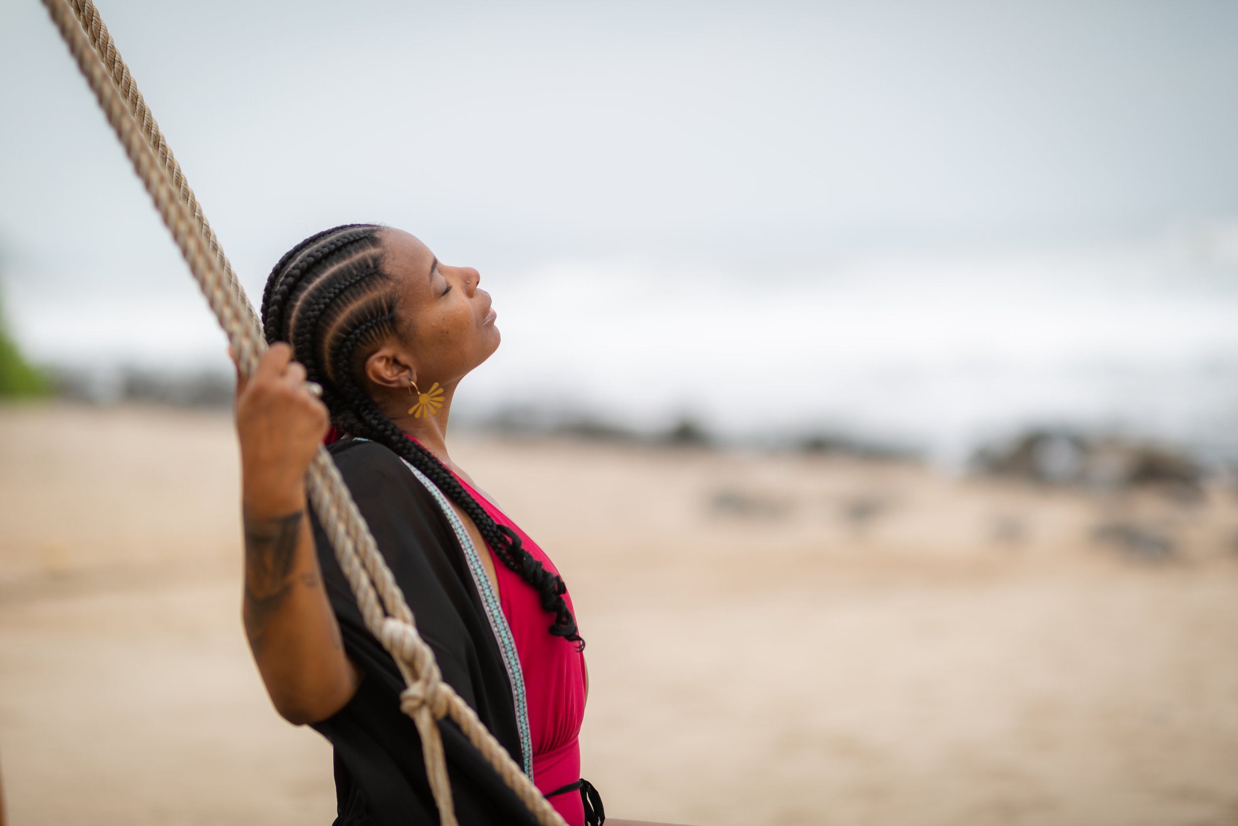 A woman with braided hair is sitting on a swing and looking up at the overcast sky on a beach.