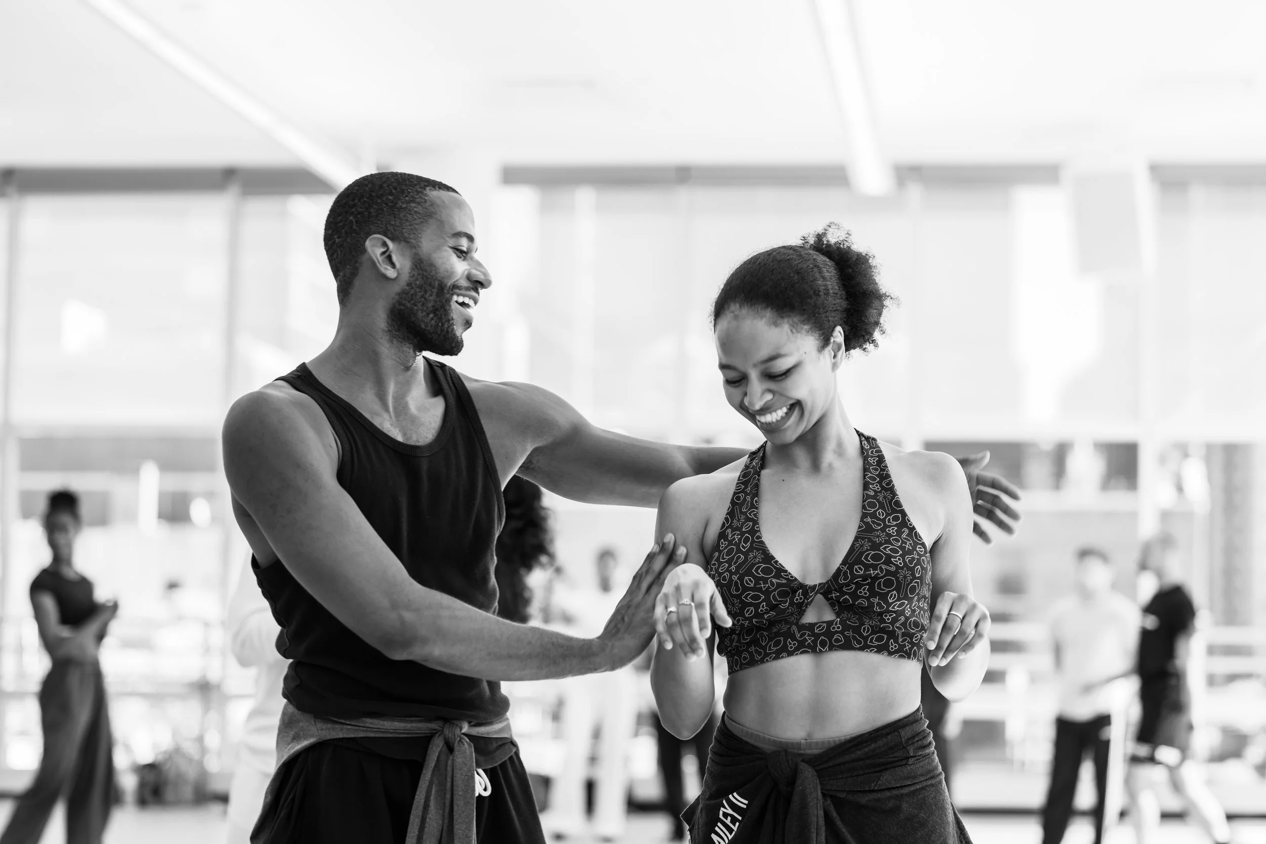 Two people, a man and a woman, smiling and interacting in a gym or fitness studio, with other individuals blurred in the background.