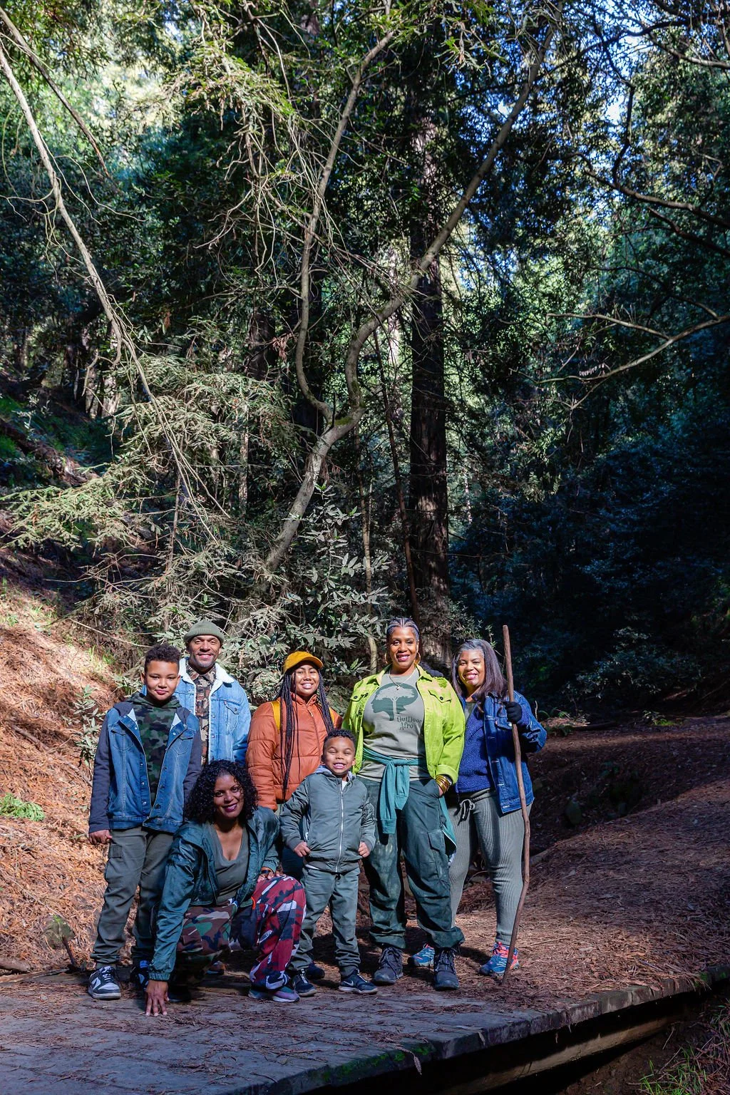 Group of seven people, diverse ages and ethnicities, standing outdoors on a trail in a forest, smiling and dressed in casual outdoor clothing with some holding walking sticks, surrounded by tall trees and dense greenery.