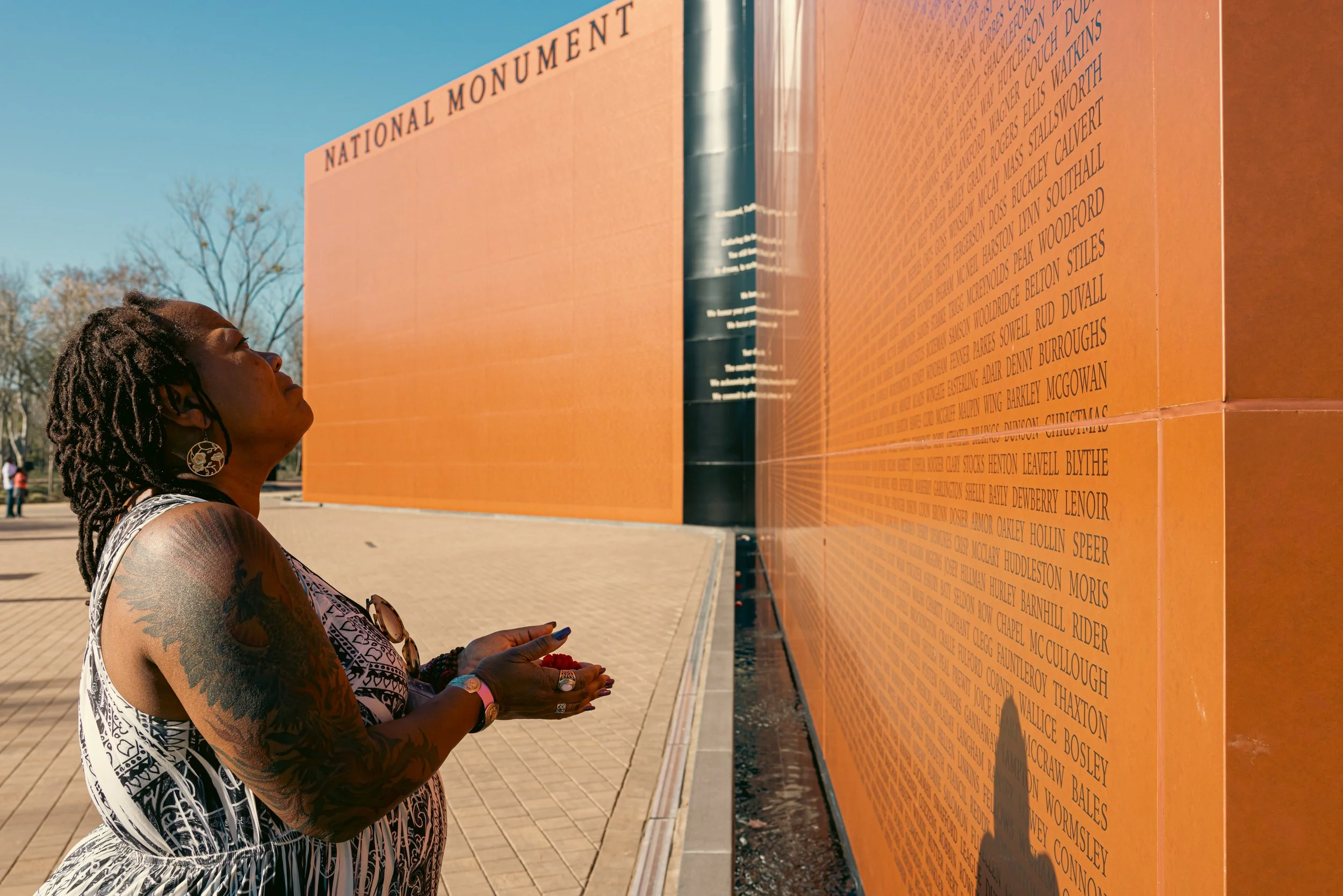 A woman with tattoos on her arm and wearing earrings looks at a bronze-colored monument wall with engraved text at the National Monument.