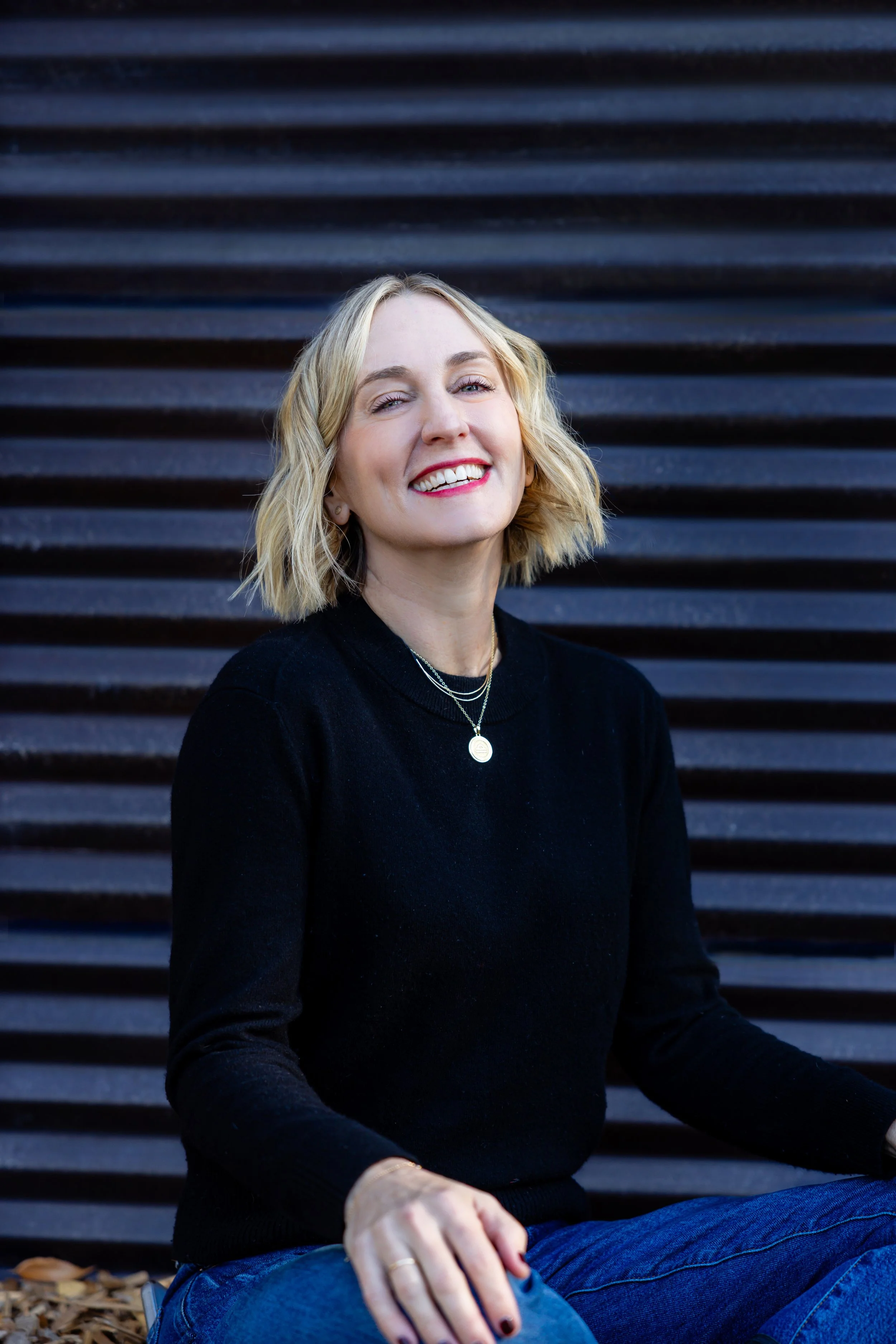 A woman with blonde, wavy hair and light skin, smiling while sitting outdoors against a black, horizontal slat background, wearing a black sweater and blue jeans.