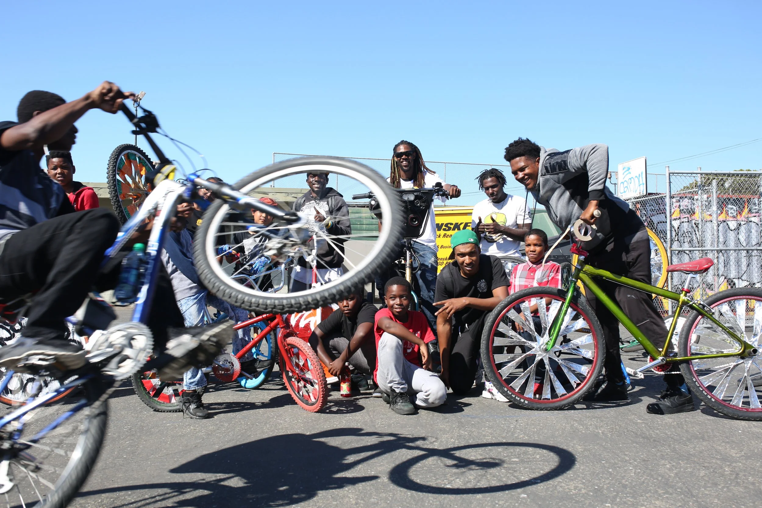 A group of people, mostly children and young adults, gathered outdoors with several colorful bicycles, some riding and some standing, on a sunny day near a chain-link fence with clear blue sky in the background.