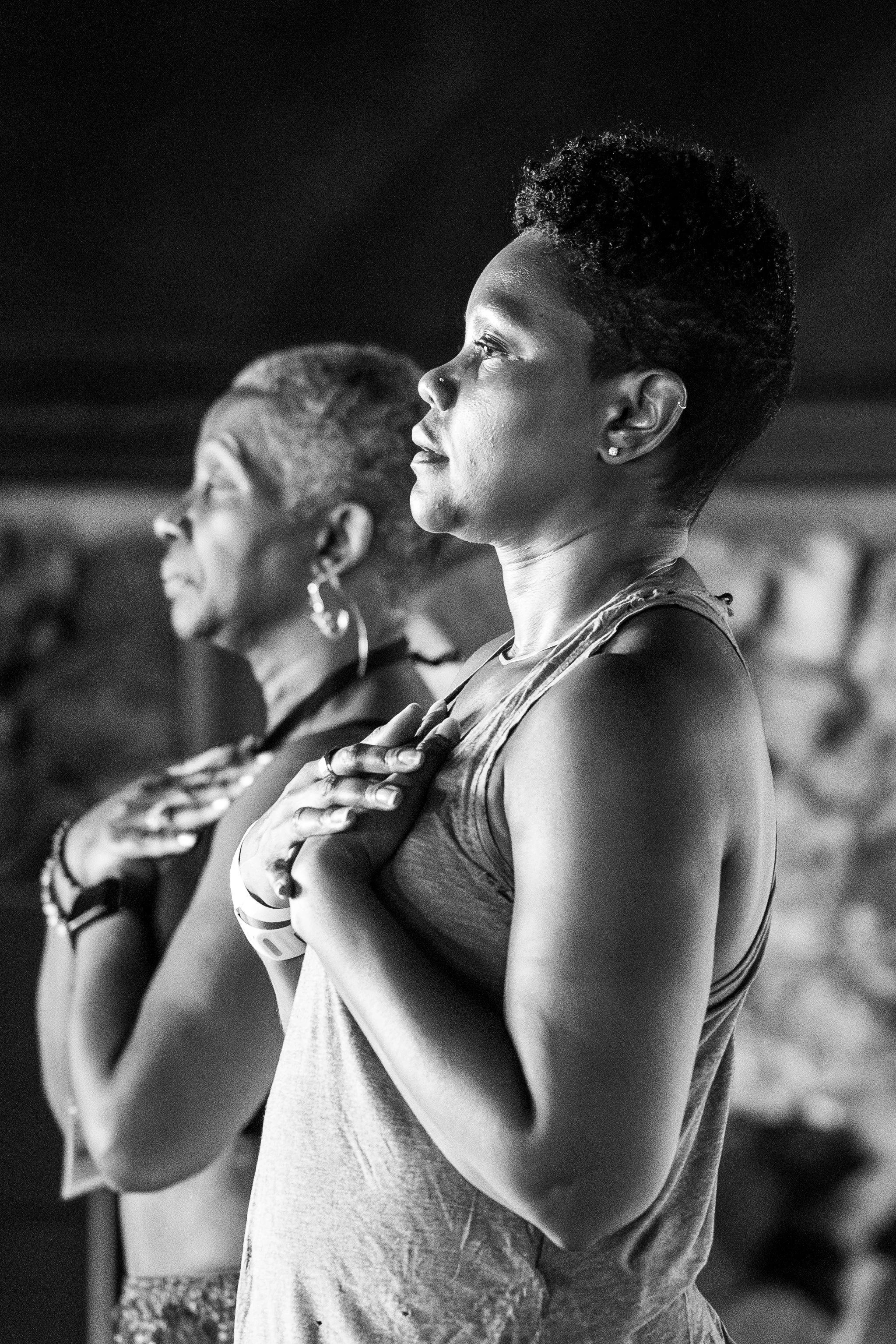 Three women with their hands over their hearts, standing side by side with eyes closed, participating in a moment of reflection or prayer, in black and white.