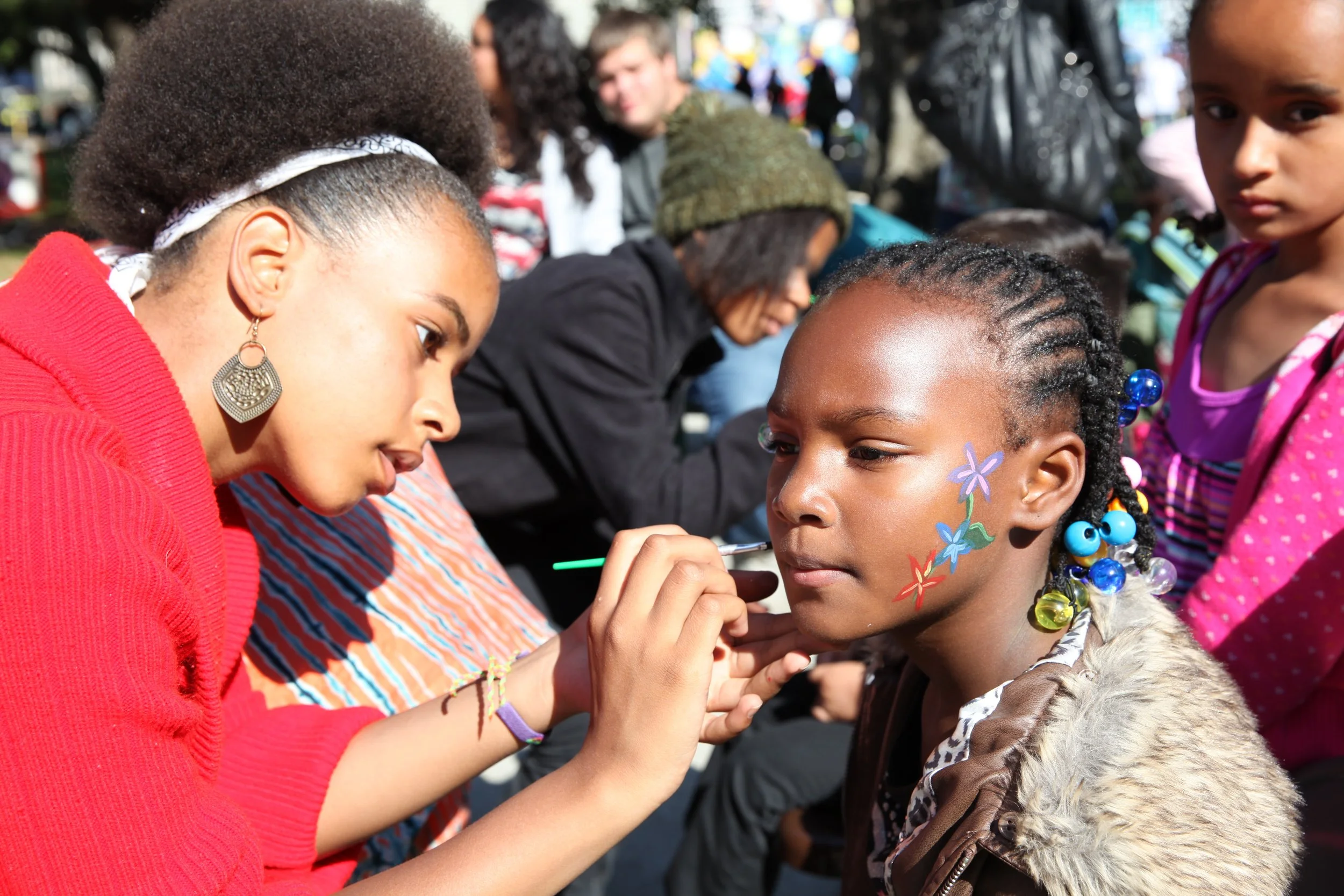 A woman is painting bright, colorful flowers on the face of a young girl with braided hair during an outdoor event. Several people are in the background, indicating a lively gathering.