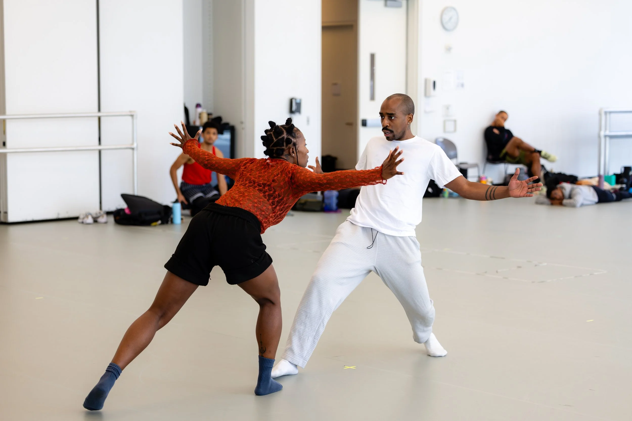 Two people practicing a martial arts move in a studio, with one person in red and black and the other in white, while others are sitting or lying on the floor in the background.