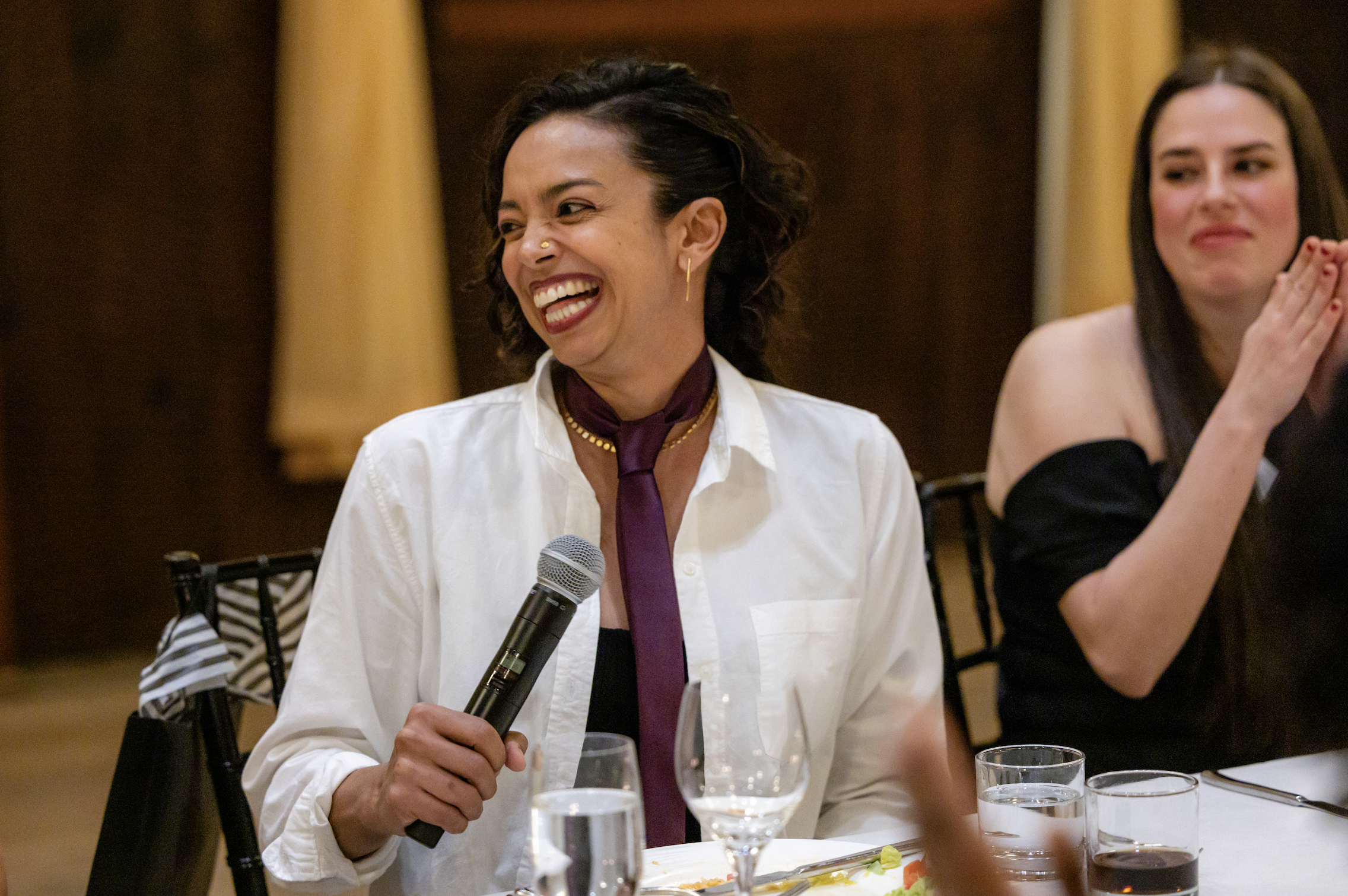 A woman with short black hair, wearing a white shirt and purple tie, is smiling and holding a microphone at a formal event. There are glasses of water and a plate of food on the table in front of her. Other people are seated nearby, and the background features yellow curtains.