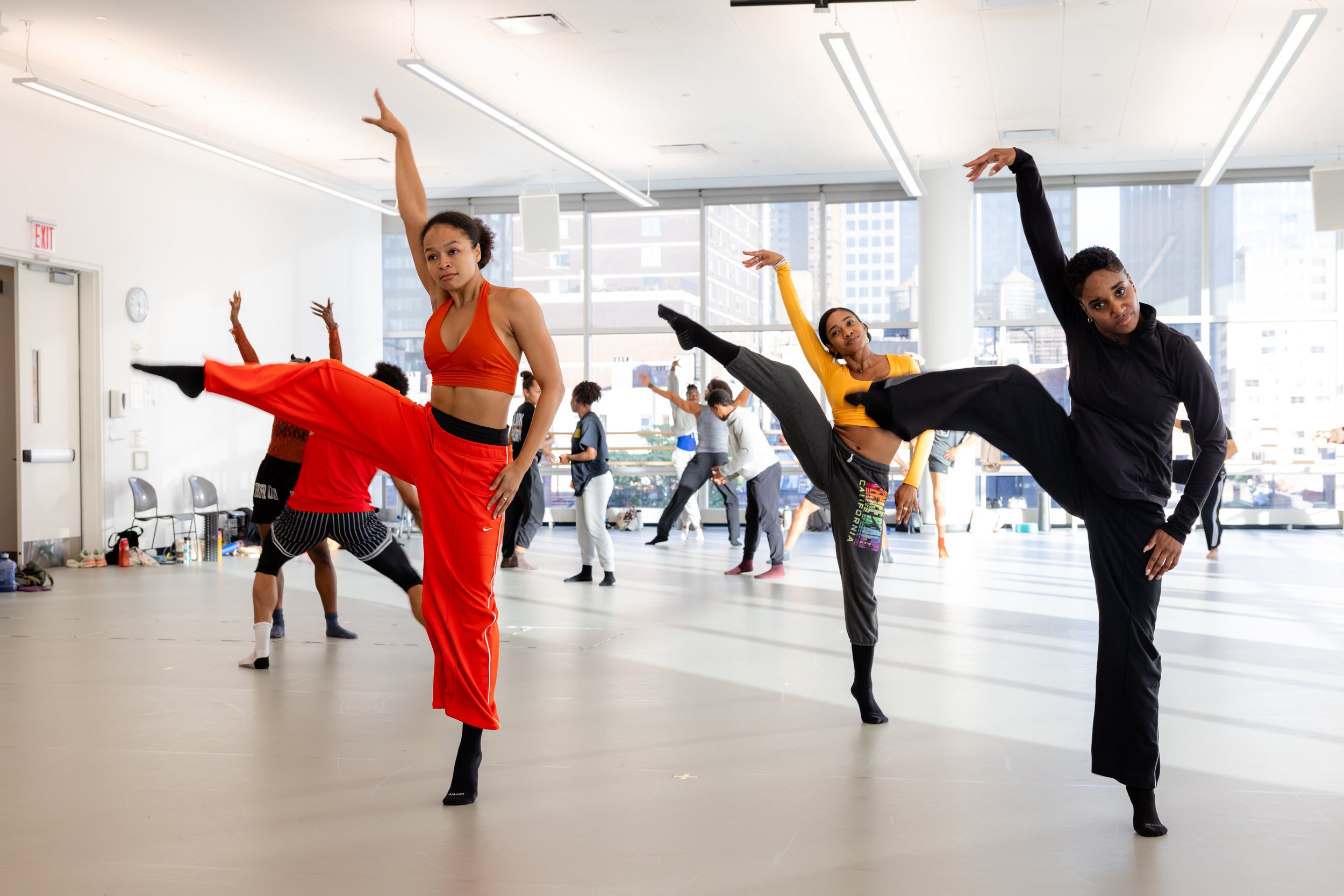 Group of diverse women practicing ballet in a dance studio with large windows, high ceilings, and city buildings outside.