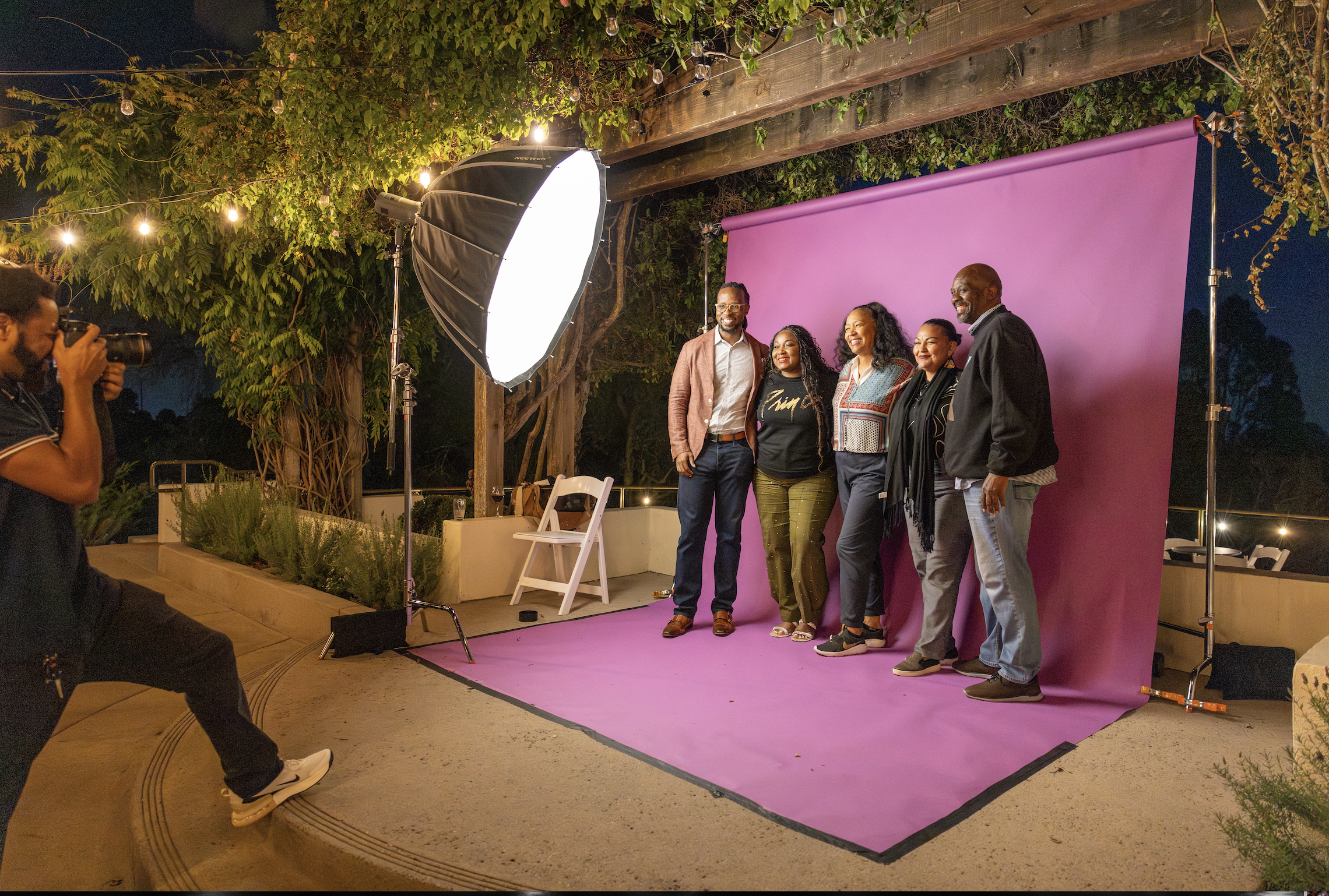 Group of five people posing for a photo in front of a pink backdrop at night, with professional lighting, outdoors under string lights and greenery.