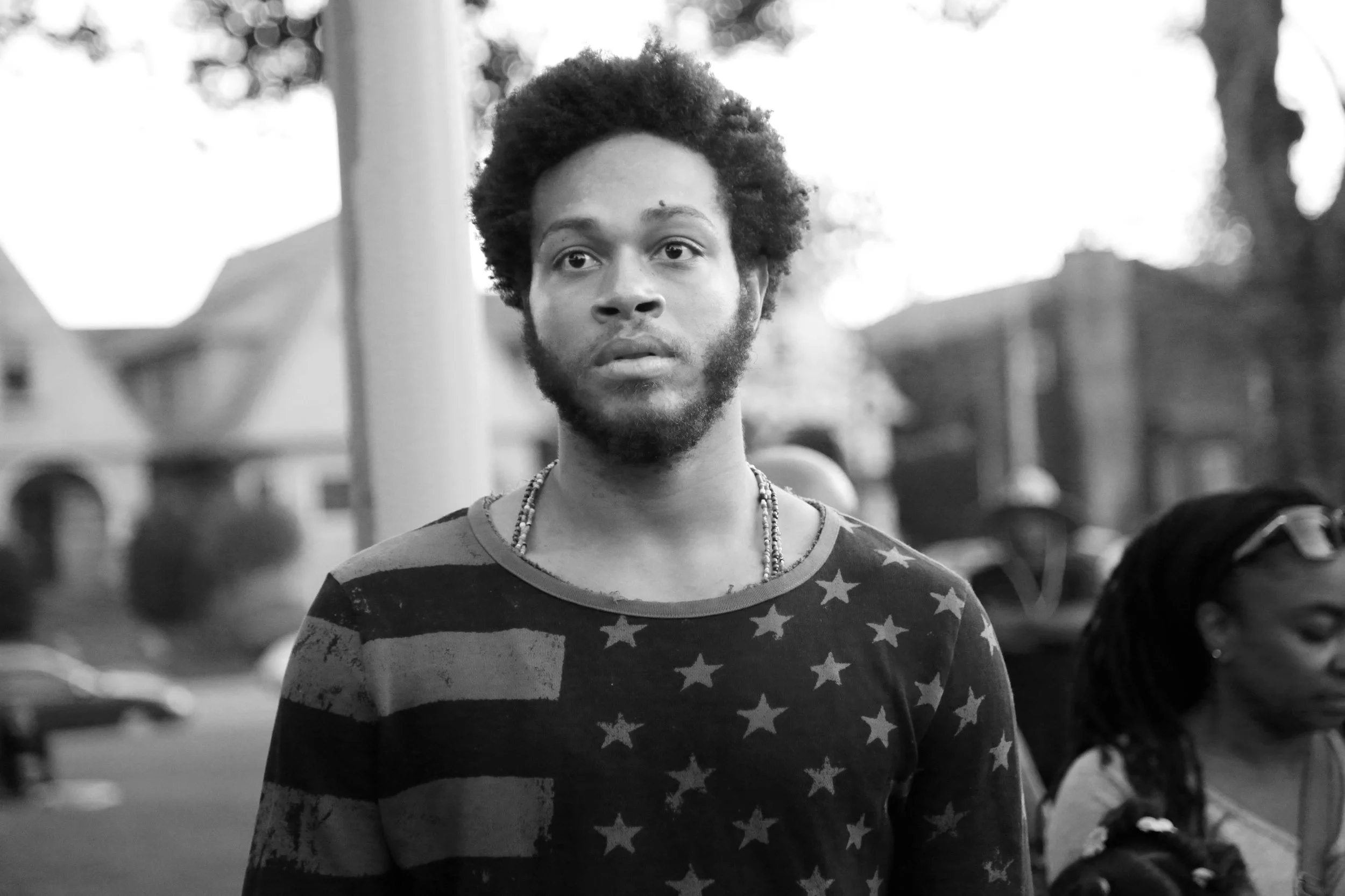 A young man with a beard and afro hairstyle wearing a USA flag shirt and necklaces, standing outdoors with trees and houses in the background, captured in black and white.