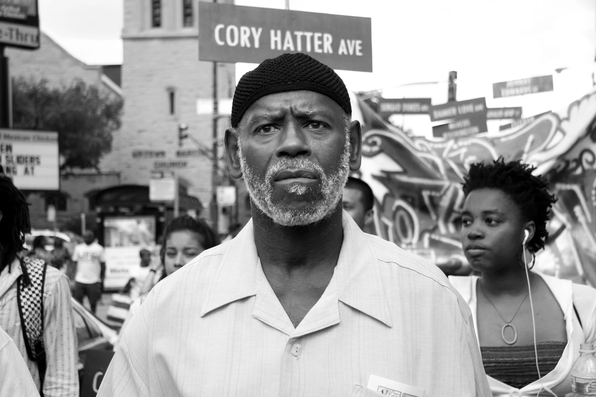 A black and white photograph of an older man with a gray beard and mustache, wearing a black knit cap and a light striped collared shirt, standing in a crowd during a protest or march. Behind him, there are several people holding signs, and a street 
