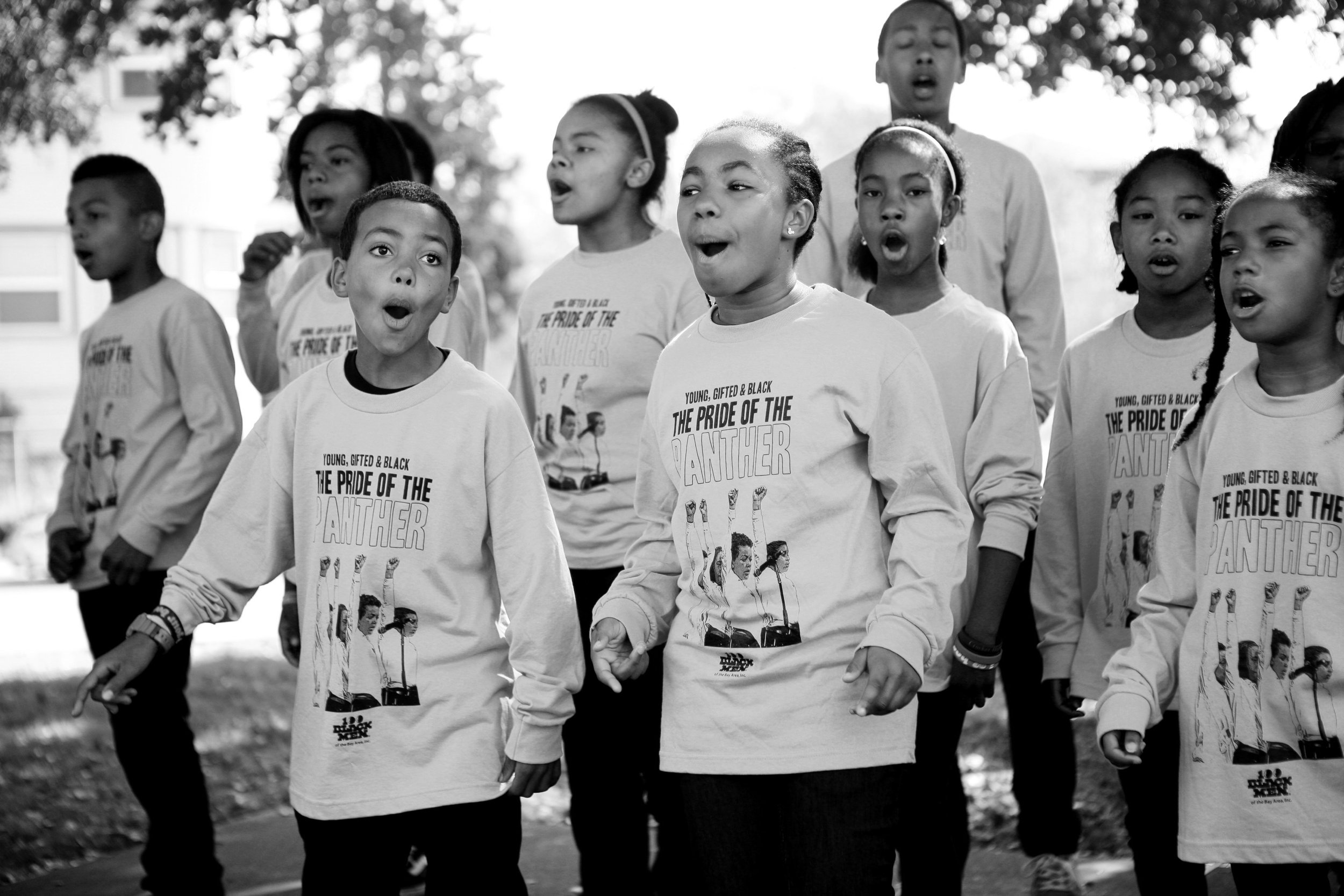 A group of children, some with their mouths open as if singing or shouting, are walking outdoors. They are wearing T-shirts with a graphic and text that says 'The Pride of the Panther' and features an image of students.