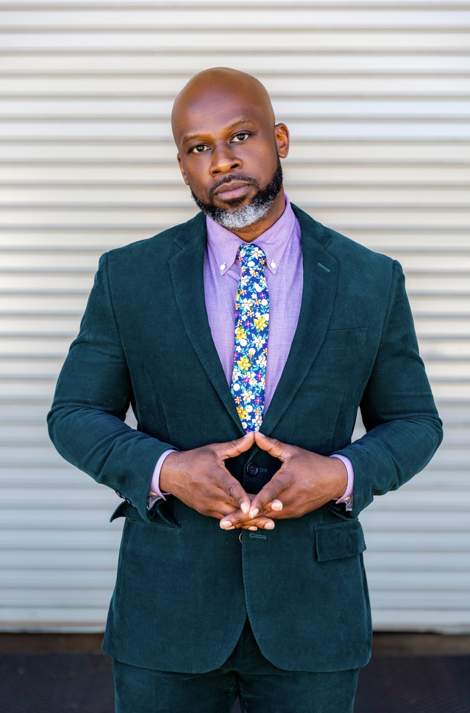 A man in a dark green blazer, purple shirt, and floral tie standing in front of a metallic horizontal slat background.
