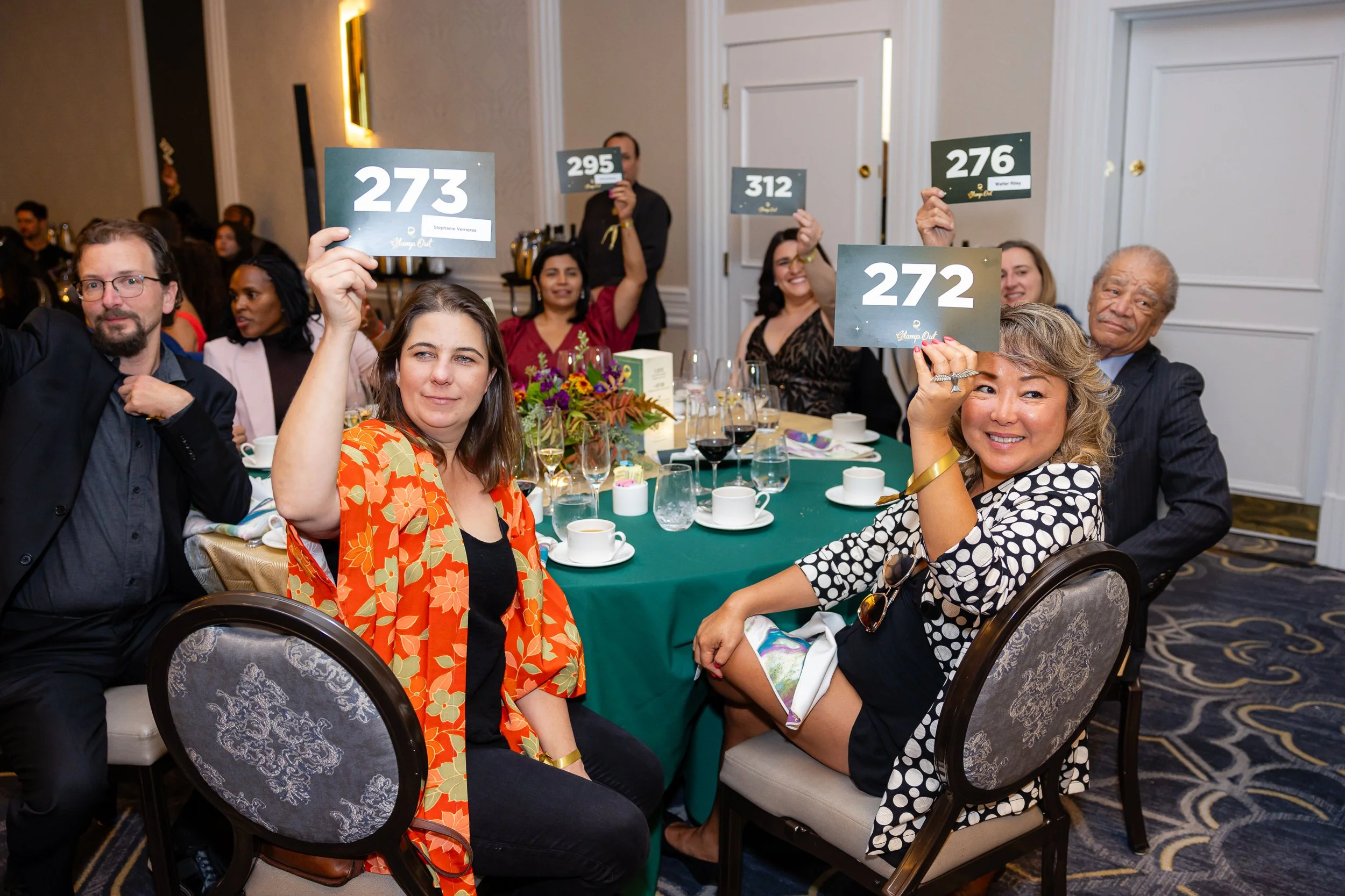 People sitting at a banquet table at an event, holding up placards with numbers, smiling and celebrating.