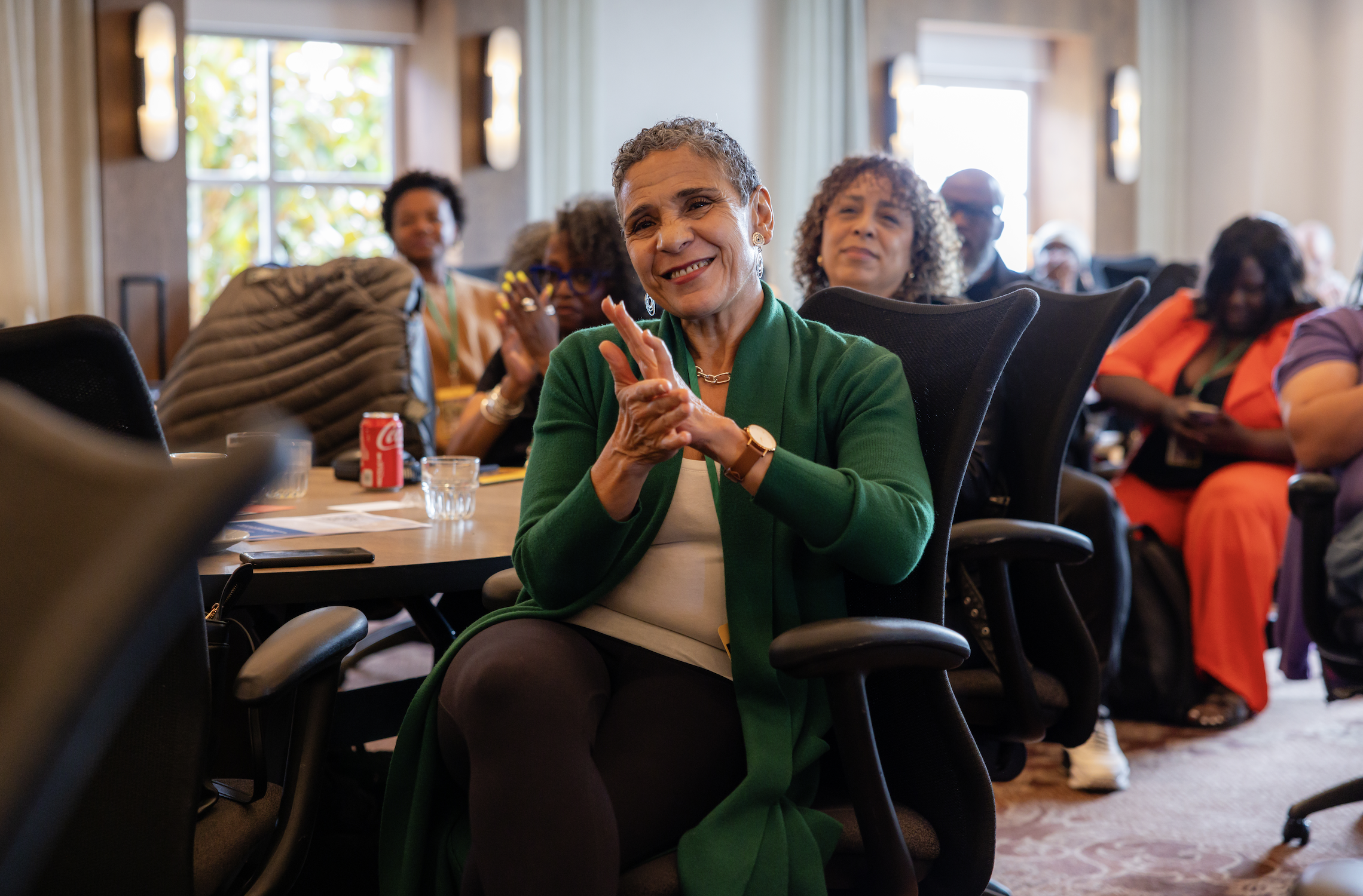 A group of diverse women in a conference room, sitting at tables, listening and clapping with smiles during a presentation.