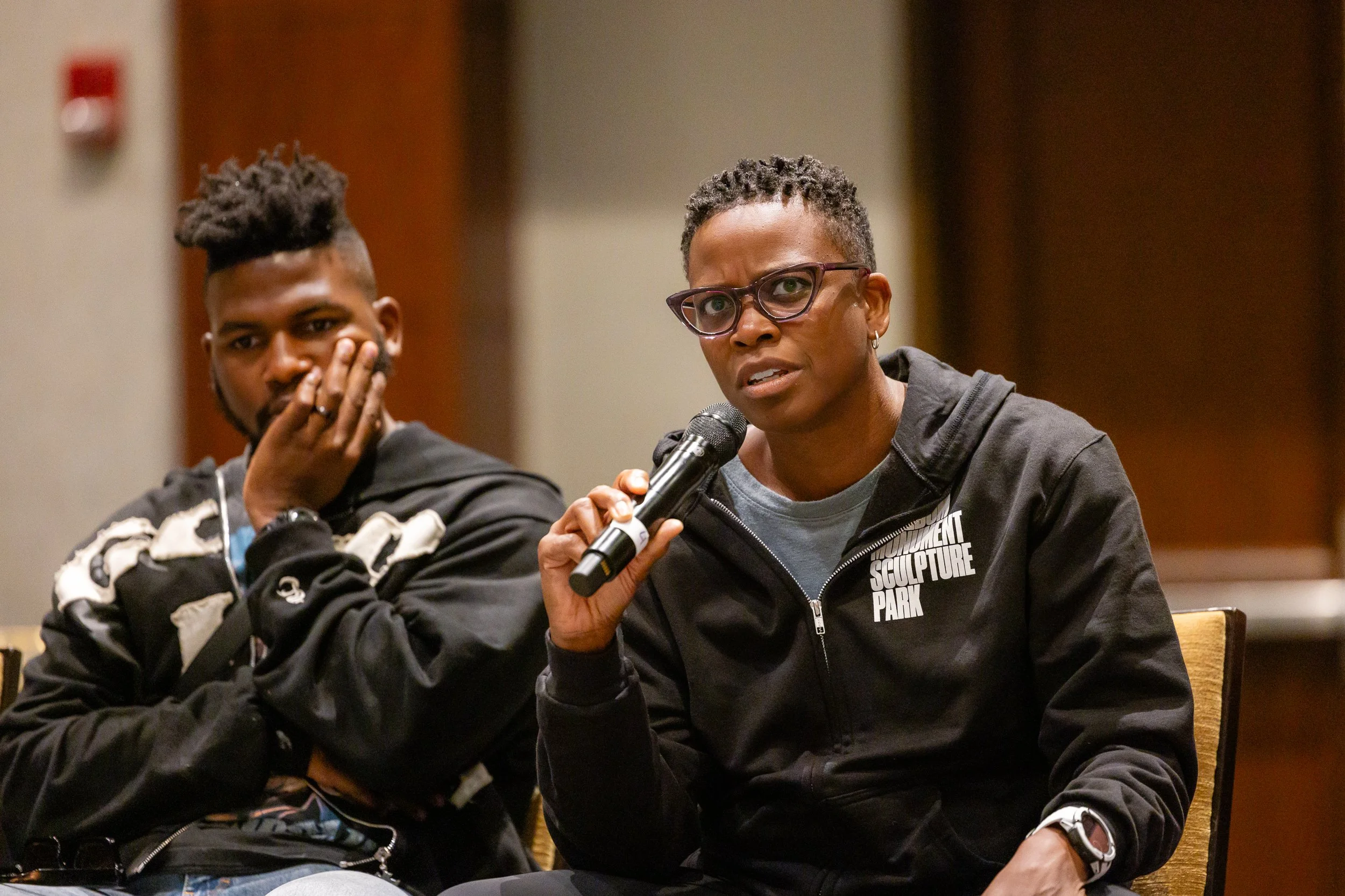 Two women seated at a panel discussion, one speaking into a microphone. The woman in the foreground has short hair, glasses, and wears a black hoodie with white text. The woman in the background has short dreadlocked hair, and is wearing a jacket wit