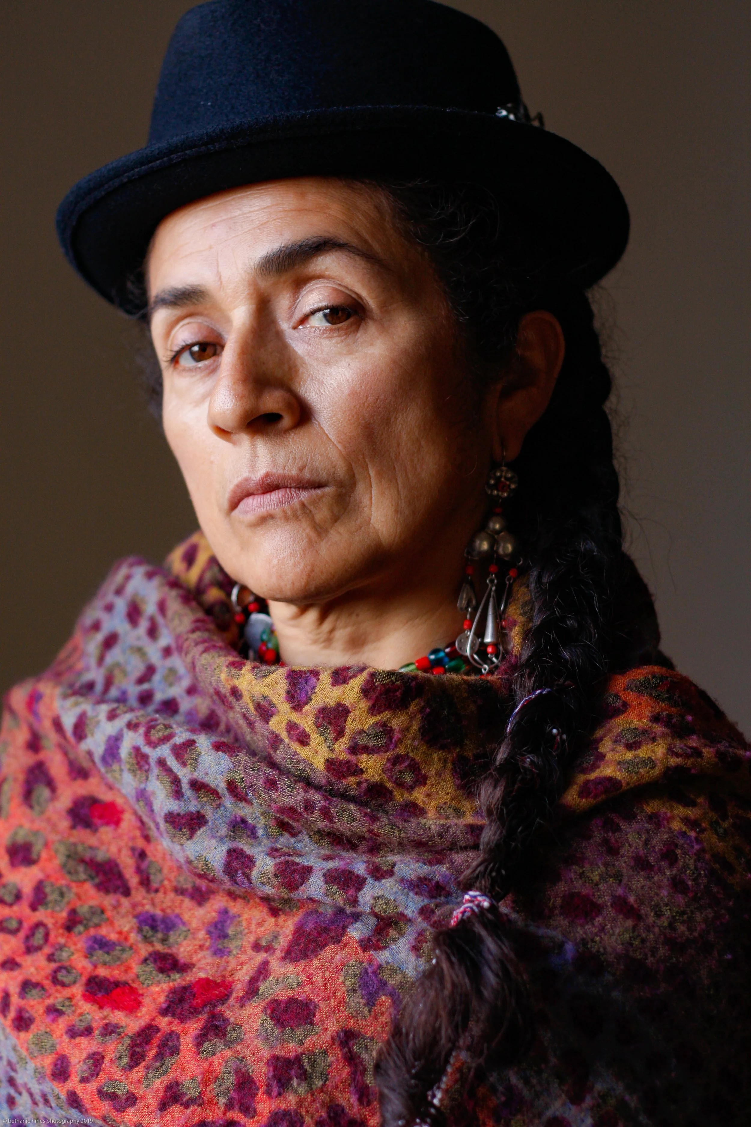 A close-up portrait of a woman with dark hair styled in a braid, wearing a black hat, colorful jewelry, and a vibrant patterned shawl.