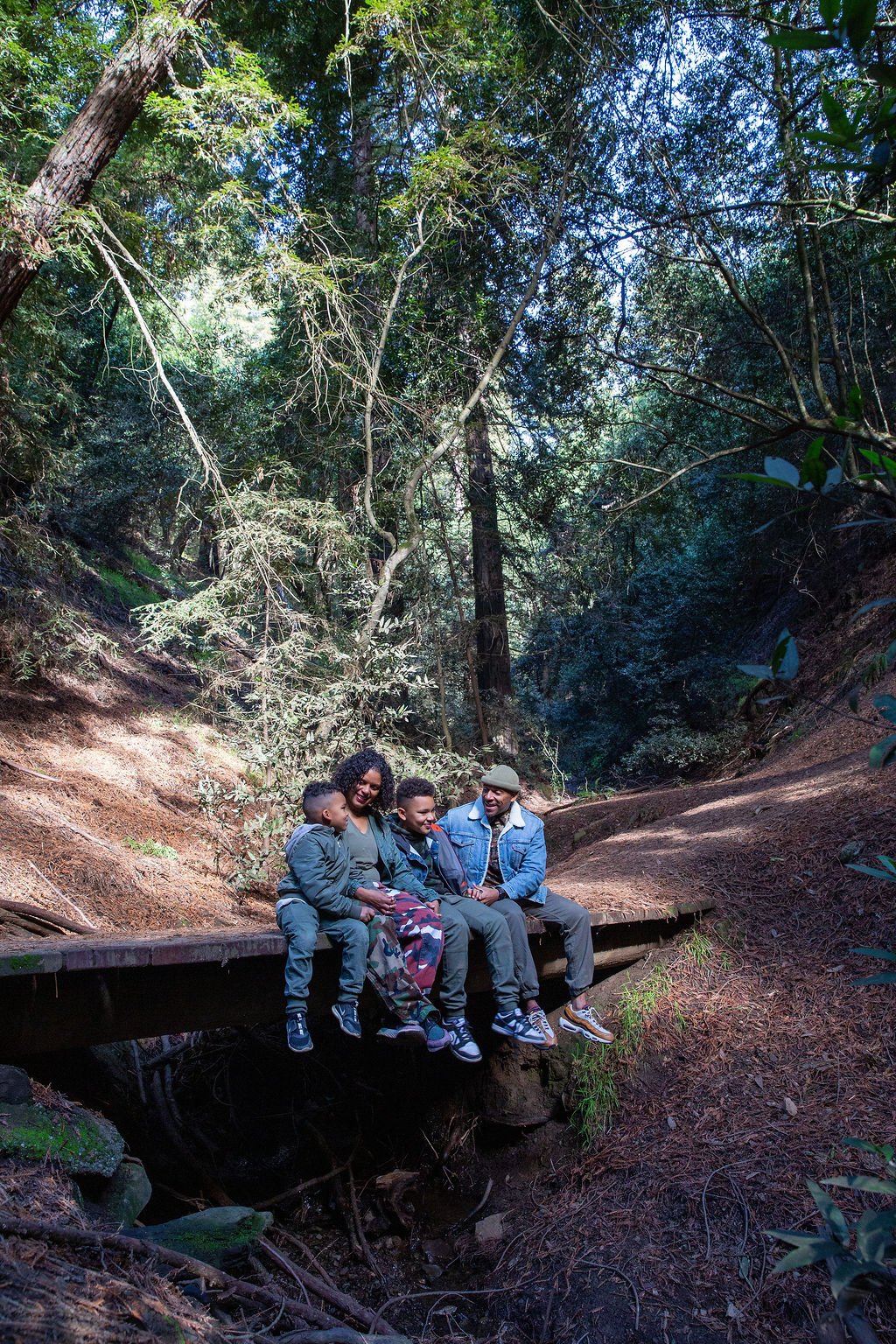 Family of four sitting on a wooden bridge in a lush forest, smiling and enjoying each other's company.