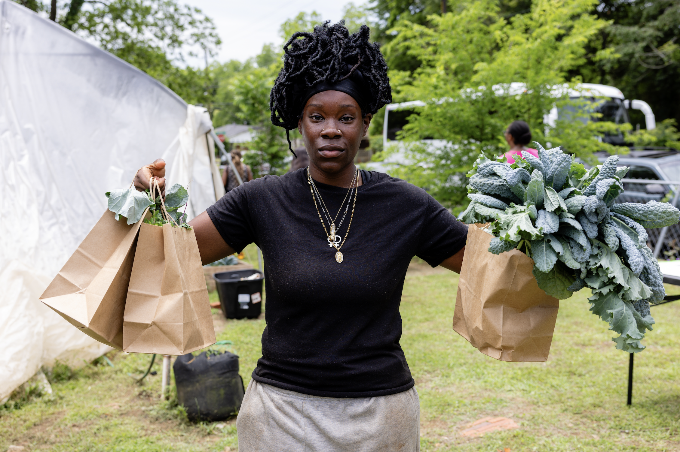Young woman outdoors holding brown paper bags filled with fresh vegetables, including leafy greens and kale, during daytime. She has dreadlocks styled in an updo and is wearing a black shirt and layered necklaces.