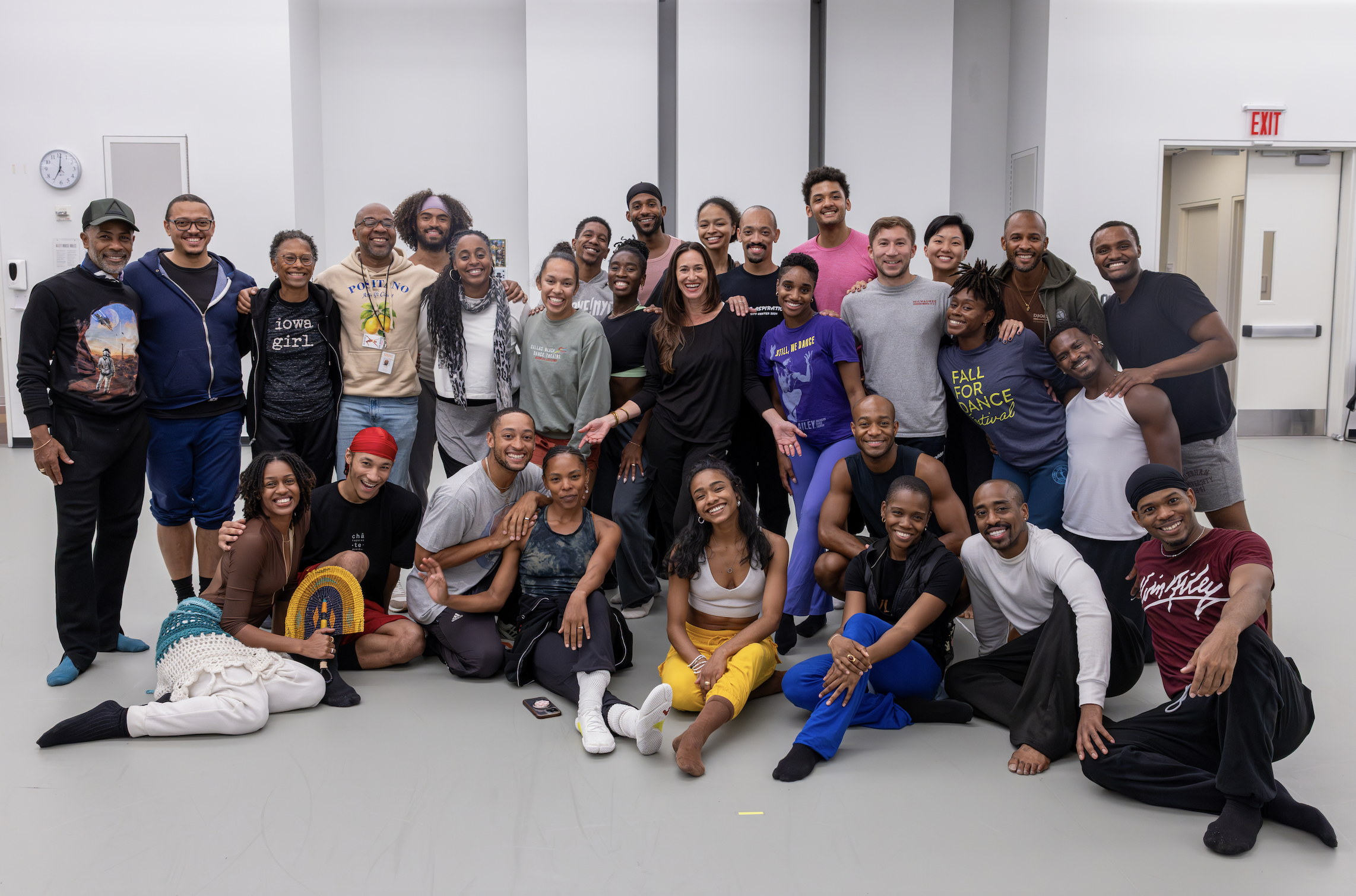 Group of diverse people smiling and posing together indoors, some sitting and some standing, with a white wall and closed door in the background.