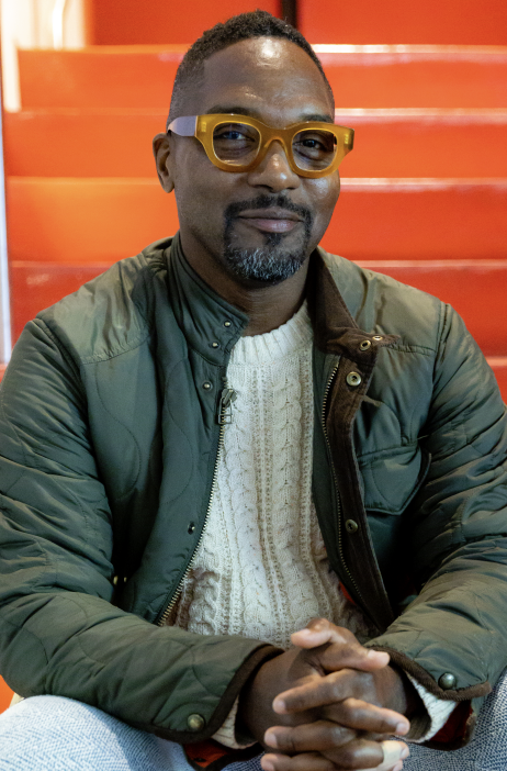 A man with dark skin, short hair, and a beard wearing yellow glasses, a green jacket, and a cream sweater, sitting in front of orange stairs.