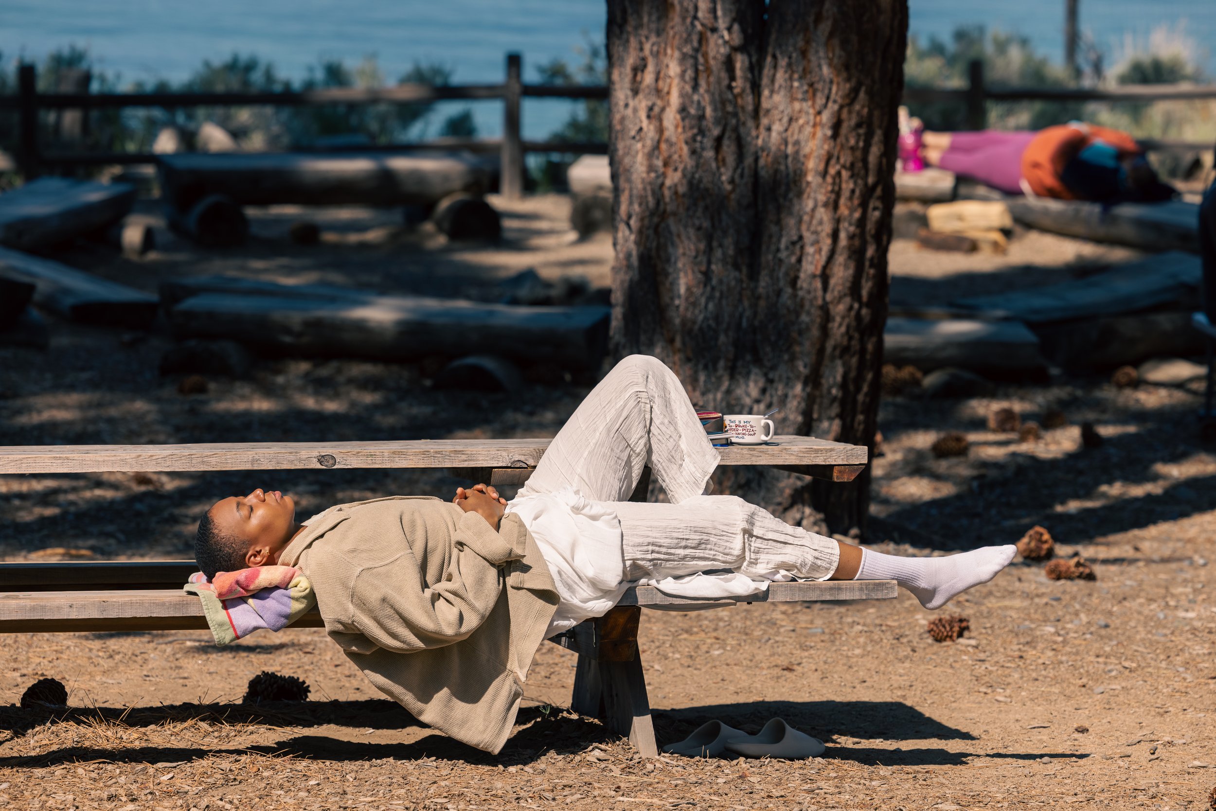 A person lying on a wooden bench outdoors near a lake, with eyes closed, wearing a beige jacket and white pants, resting under a tree with a blanket and belongings nearby.