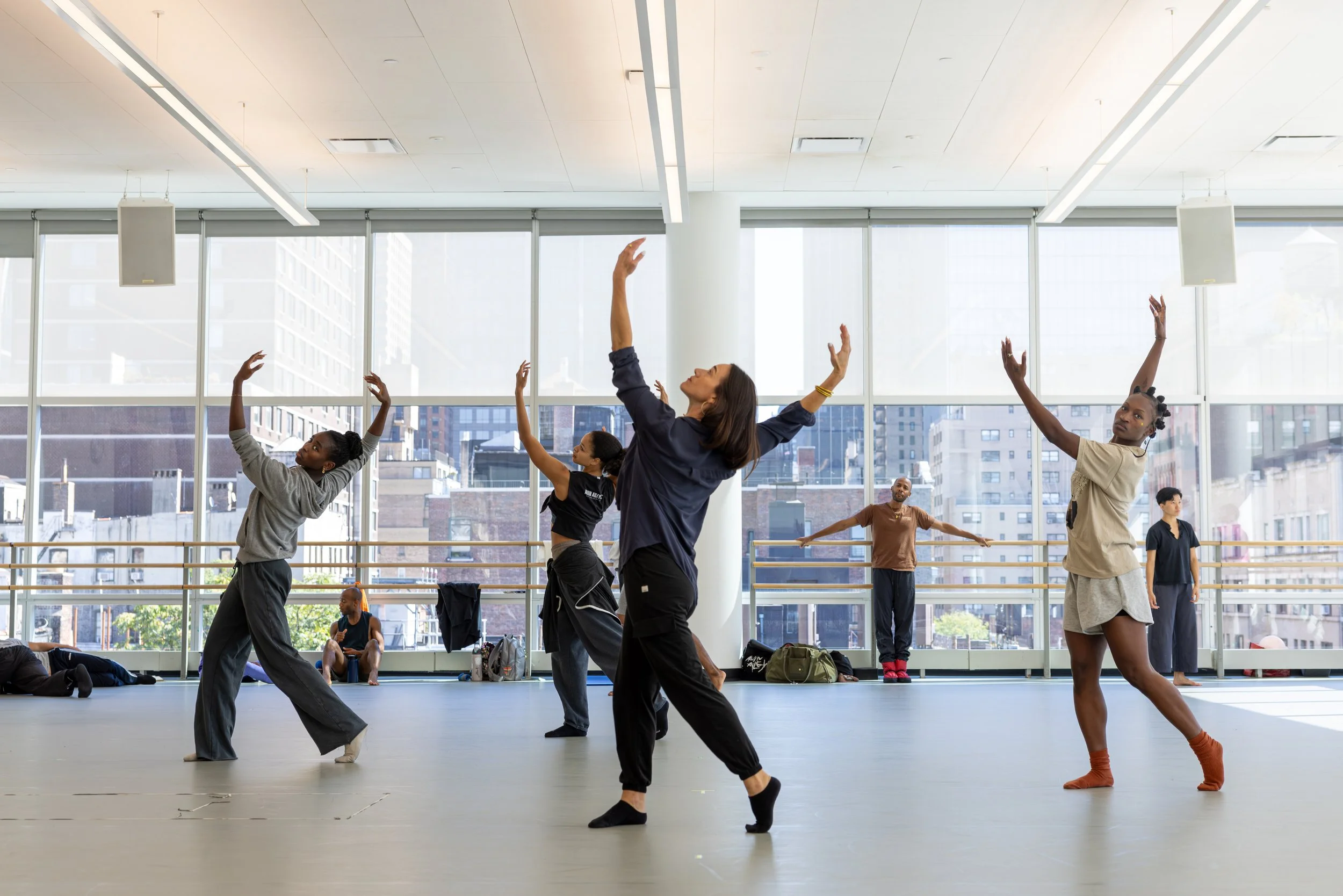 Group of diverse dancers practicing ballet in a bright, spacious studio with large windows showing city buildings outside.