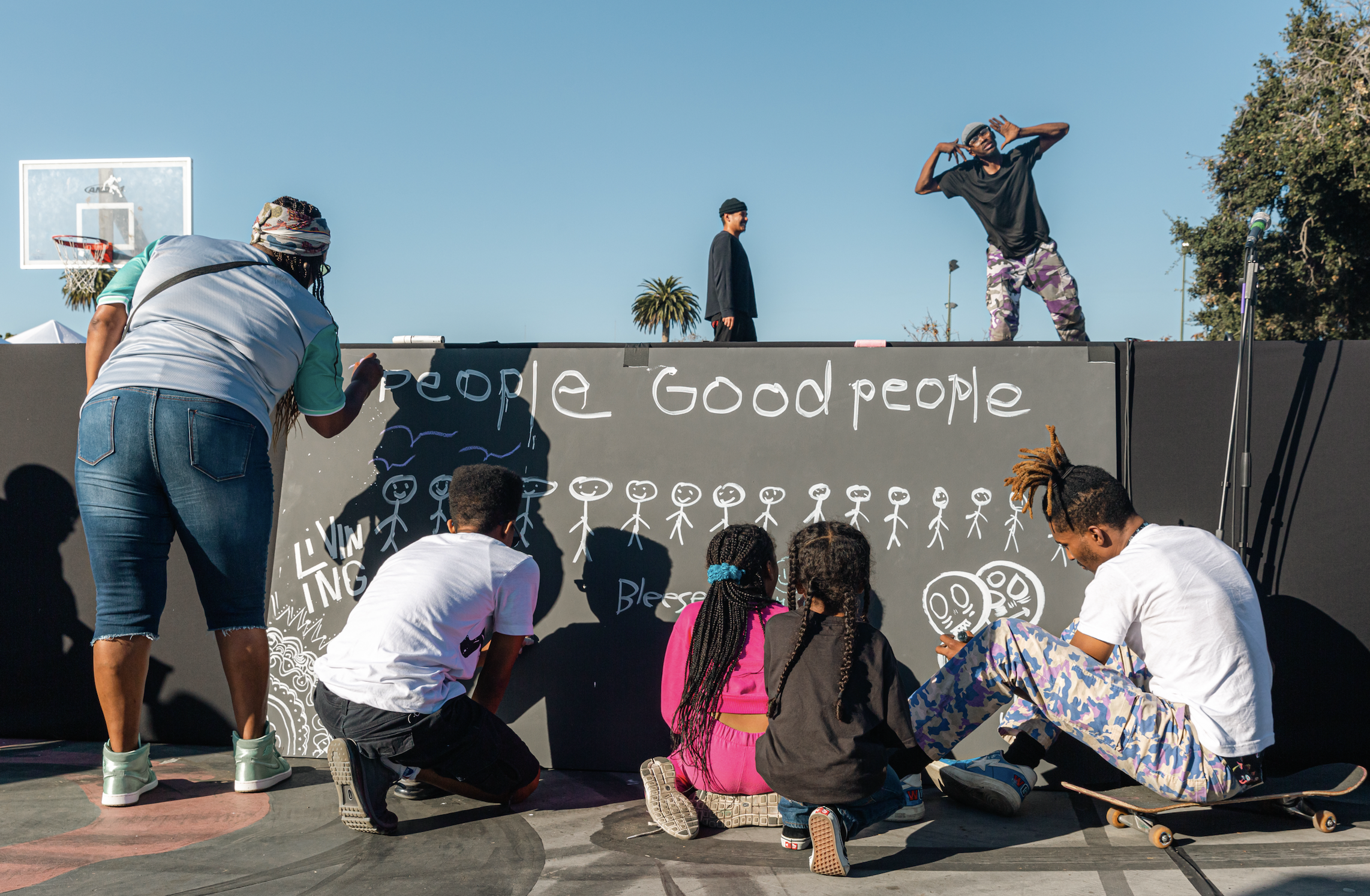 A group of children and a woman gathered in front of a blackboard at an outdoor skateboarding event. The woman is sketching on the blackboard and the children are watching or drawing. Two performers are on a raised platform, one with a hat and sungla