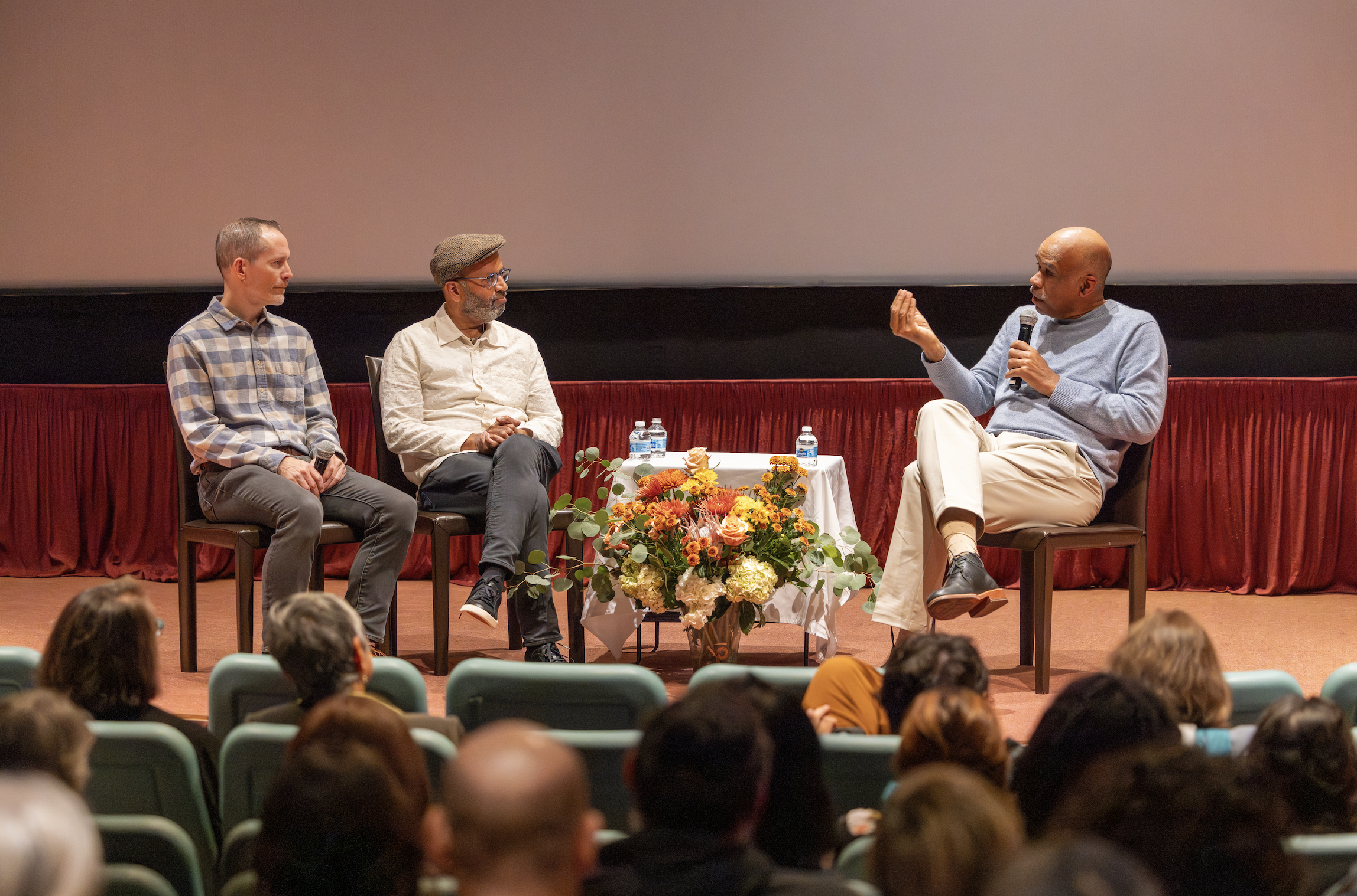 A panel discussion on stage with three men sitting on chairs, one holding a microphone, with an audience in front and a large blank screen behind them. There is a table with water bottles and a flower arrangement in front of the panel.