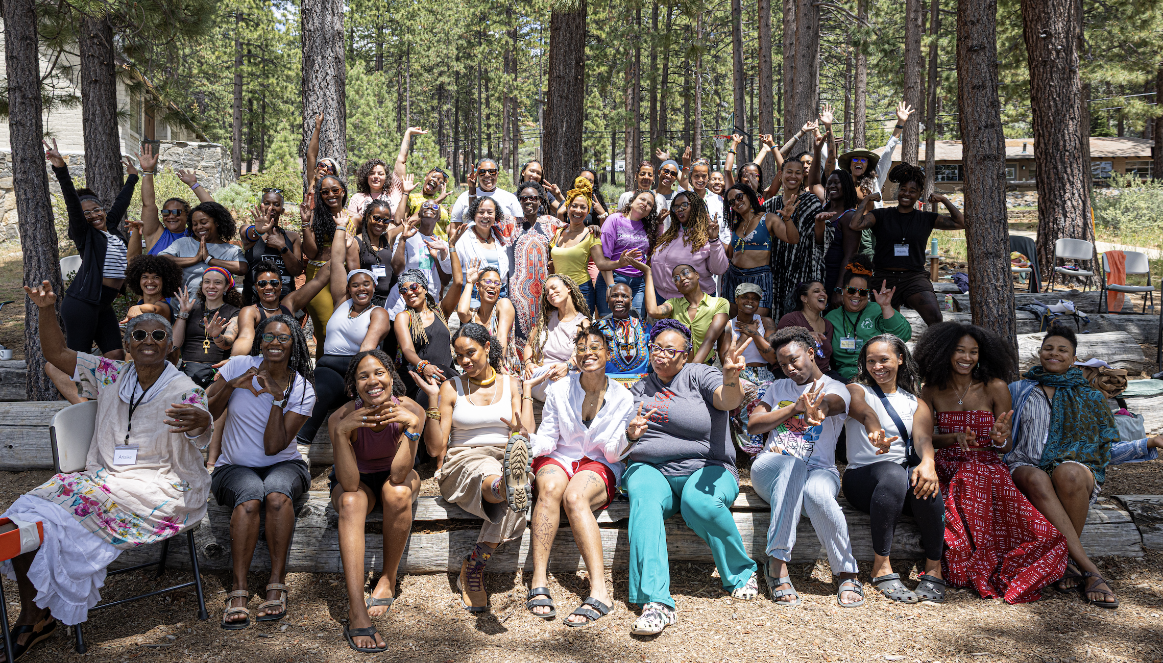 A large group of women gathered outdoors in a forested area, smiling and posing for a group photo during daytime, surrounded by tall trees.