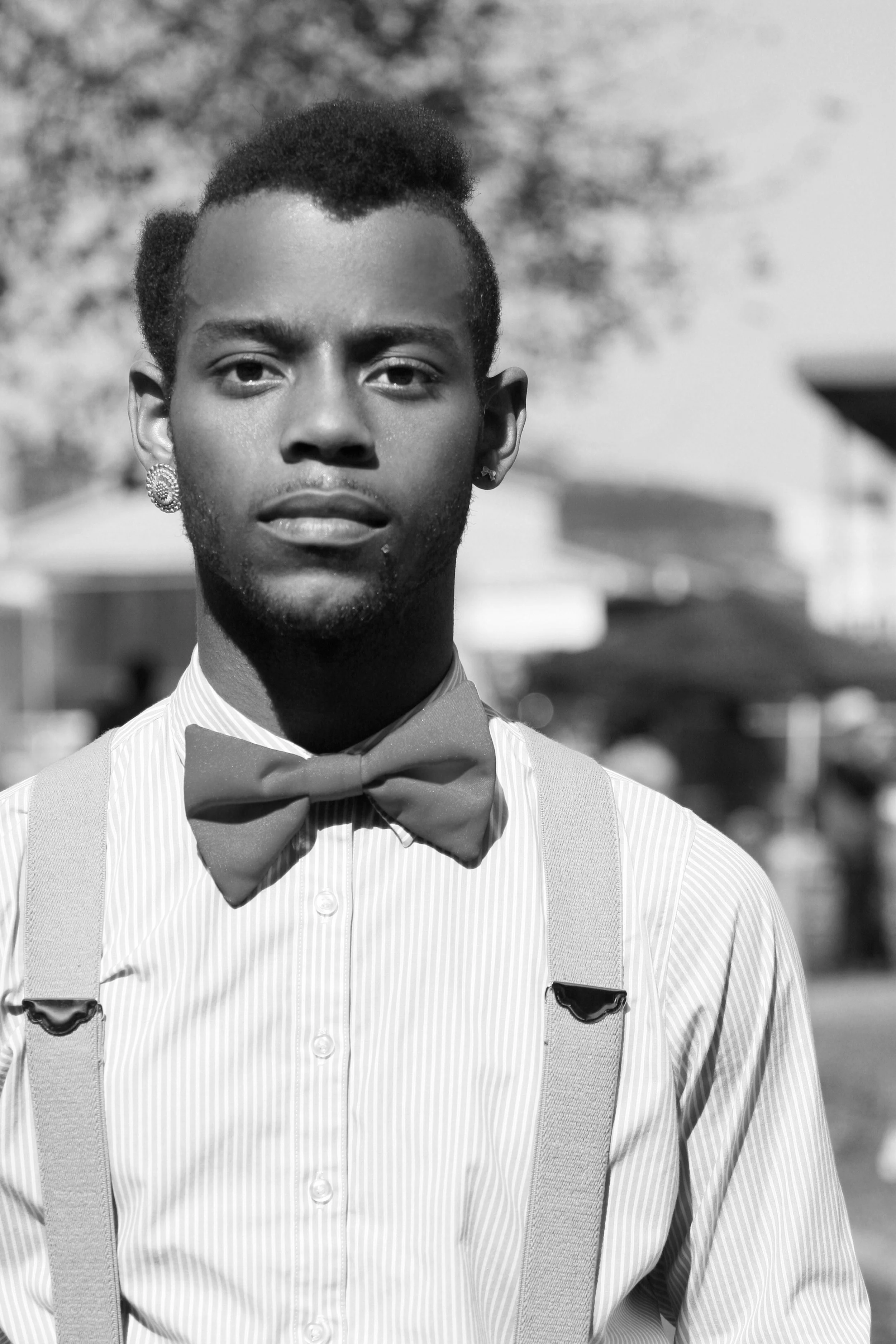 A young man with short hair, wearing a striped dress shirt, bow tie, suspenders, and earrings standing outdoors. The background is blurred with trees and sky.