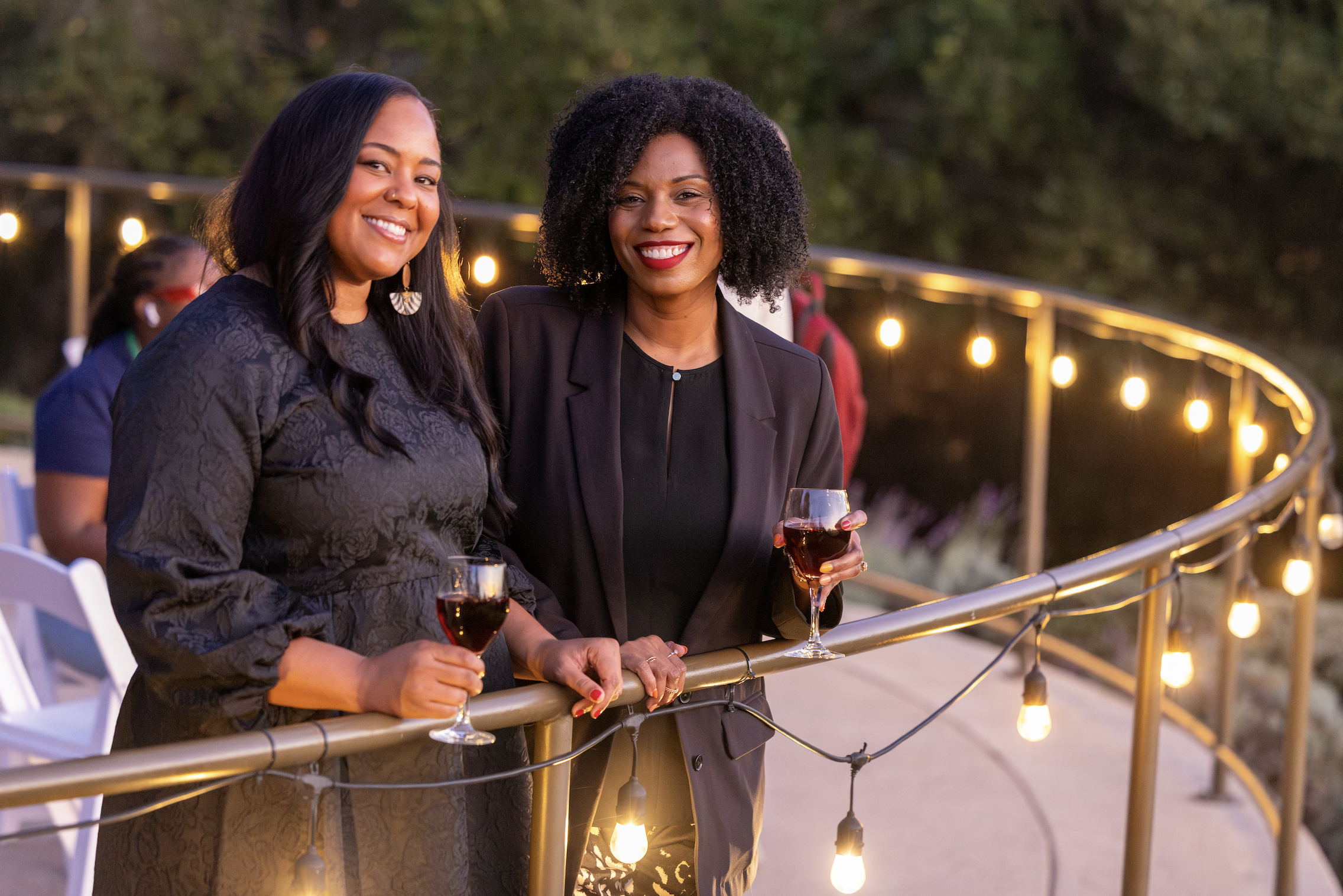 Two women smiling and holding glasses of red wine at an outdoor evening event, standing by a lit railing with string lights in the background.