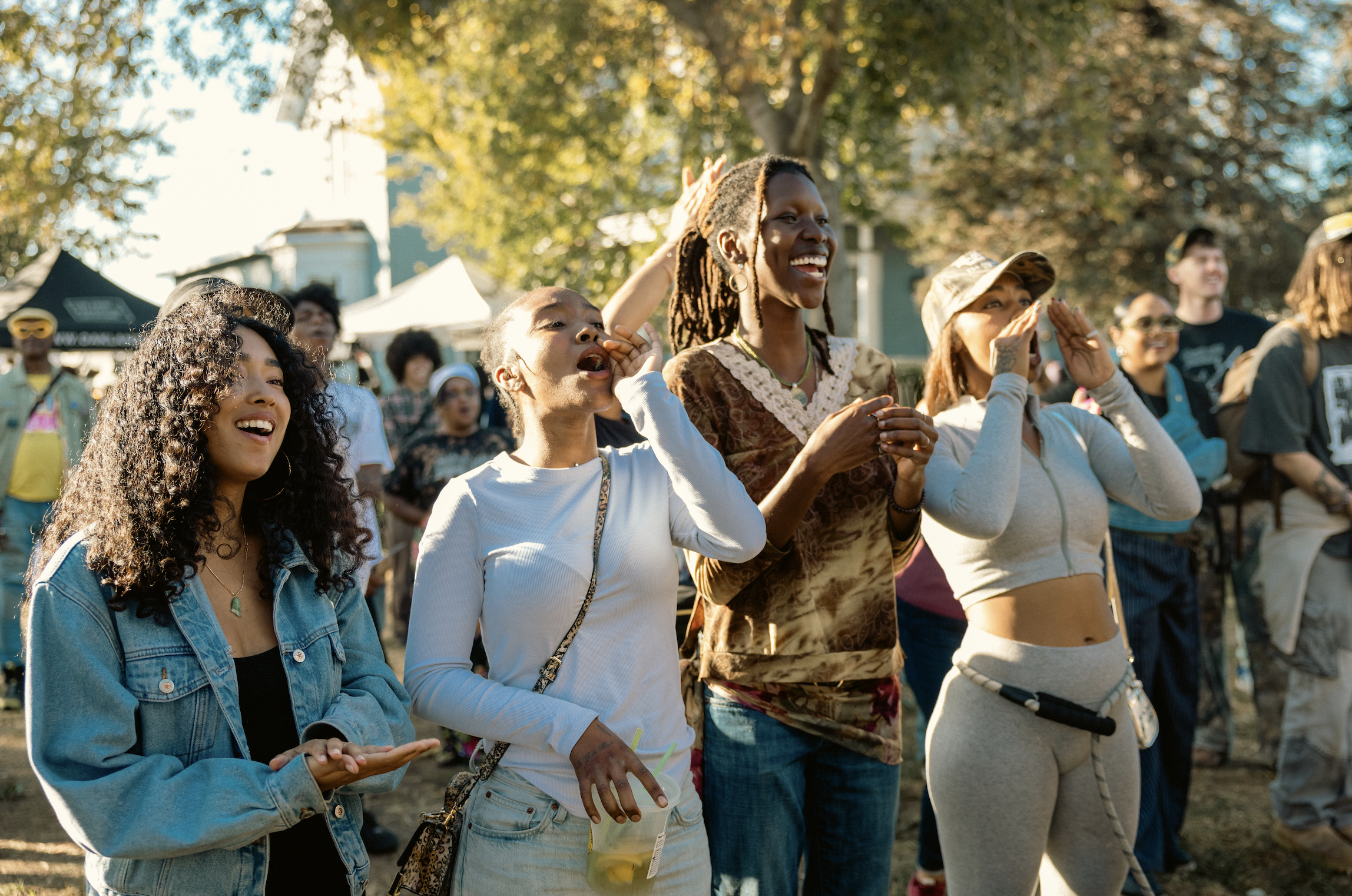 A group of diverse women smiling and cheering at an outdoor event on a sunny day, with trees and tents in the background.