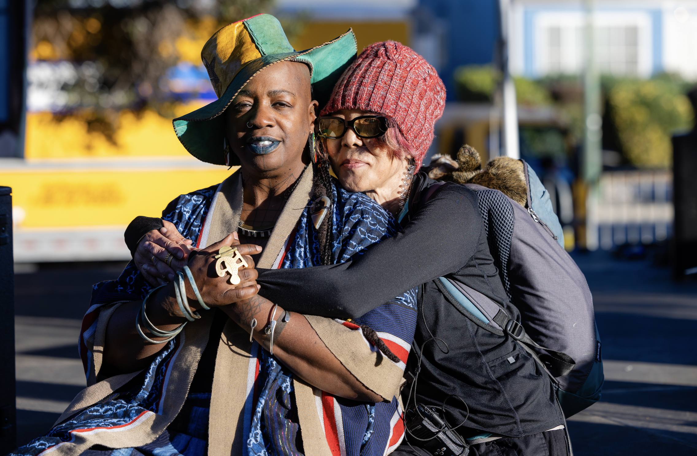 Two women hugging on a city street, wearing colorful and unique hats, with a blurred background of yellow vehicle and green trees.