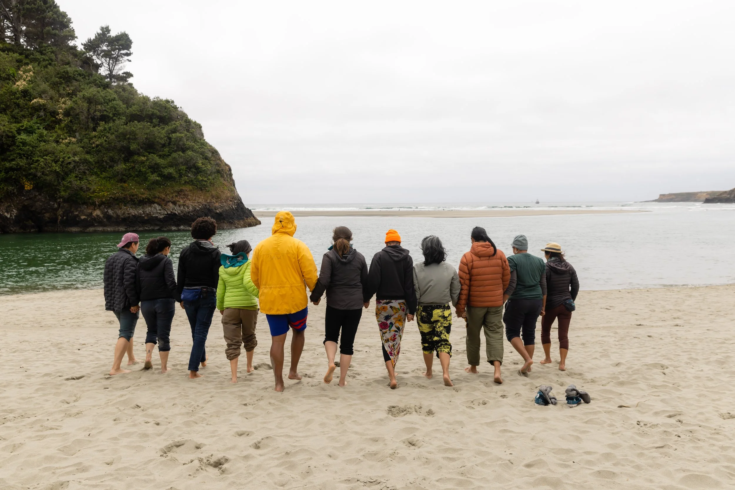 A group of people, dressed warmly, walking together barefoot on a sandy beach, with water and cliffs in the background.