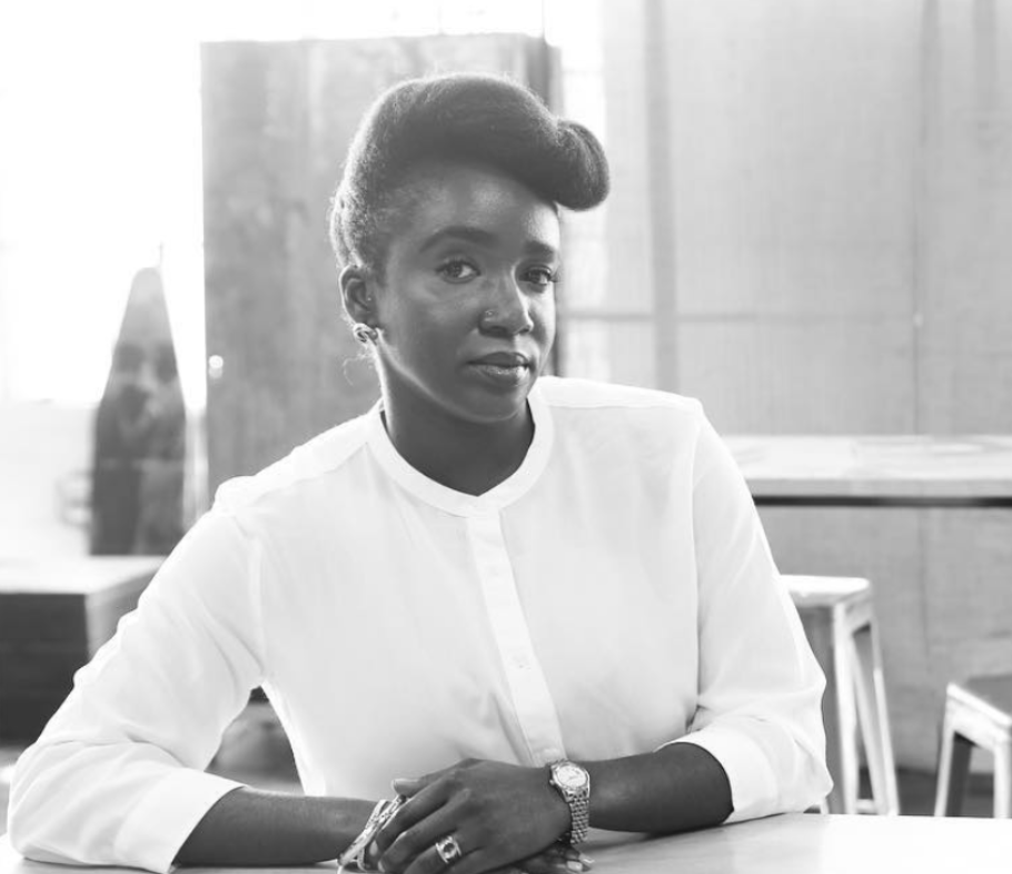 A woman with styled hair and wearing a white blouse sitting at a table in a bright room.