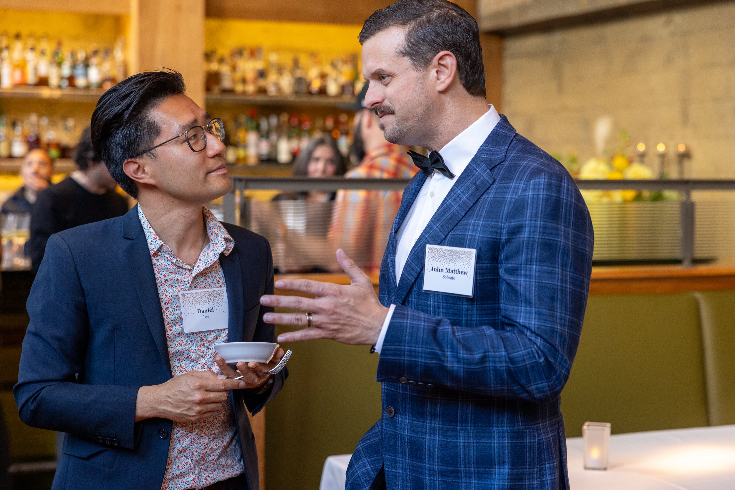 Two men engaged in conversation at a social event, one wearing glasses and a dark suit with a floral shirt, holding a small bowl, and the other in a blue checkered suit with a black bow tie, wearing a name tag that reads 'John Matthew Sobrado'. Background includes a bar with bottles and other people mingling in a warmly lit setting.