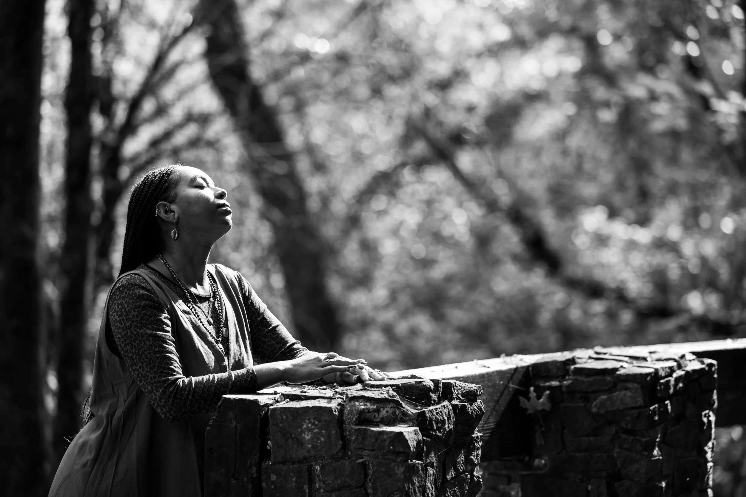 A woman with braided hair and earrings, dressed in patterned clothing, stands outdoors with her eyes closed, facing sunlight, in front of a stone ledge and blurred trees in the background.