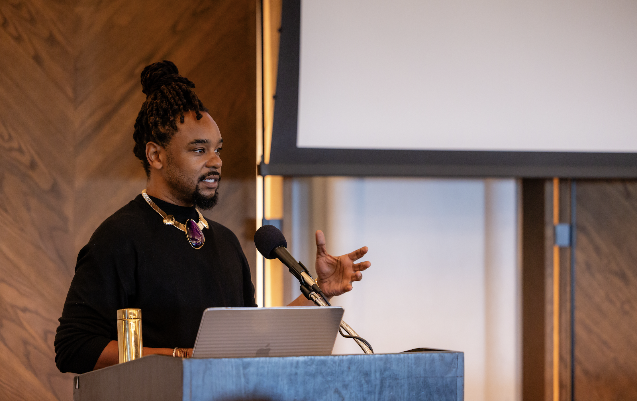 A man with dreadlocks and a goatee speaking at a podium during a presentation. He is wearing a black shirt, a statement necklace, and has a microphone in front of him.