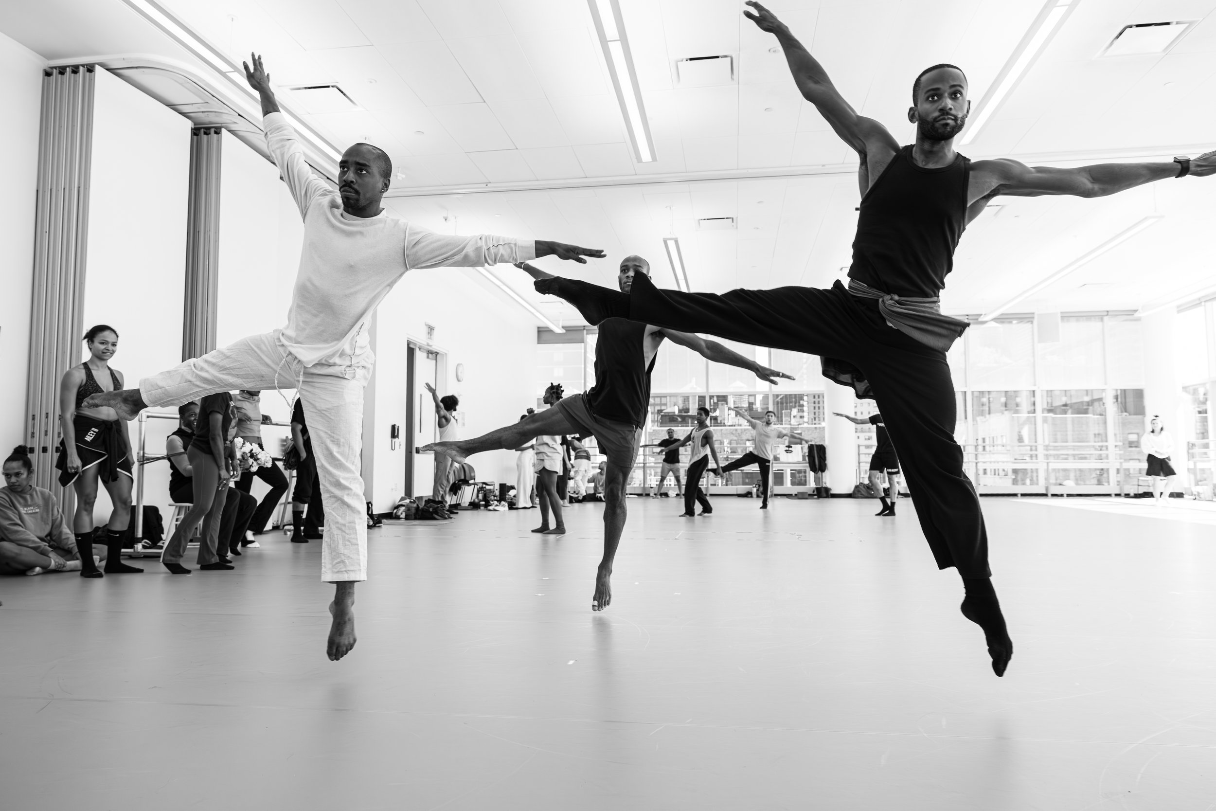 Group of ballet dancers practicing in a dance studio, with a man in a black tank top and loose pants in the foreground performing an arabesque, surrounded by other dancers stretching or standing.