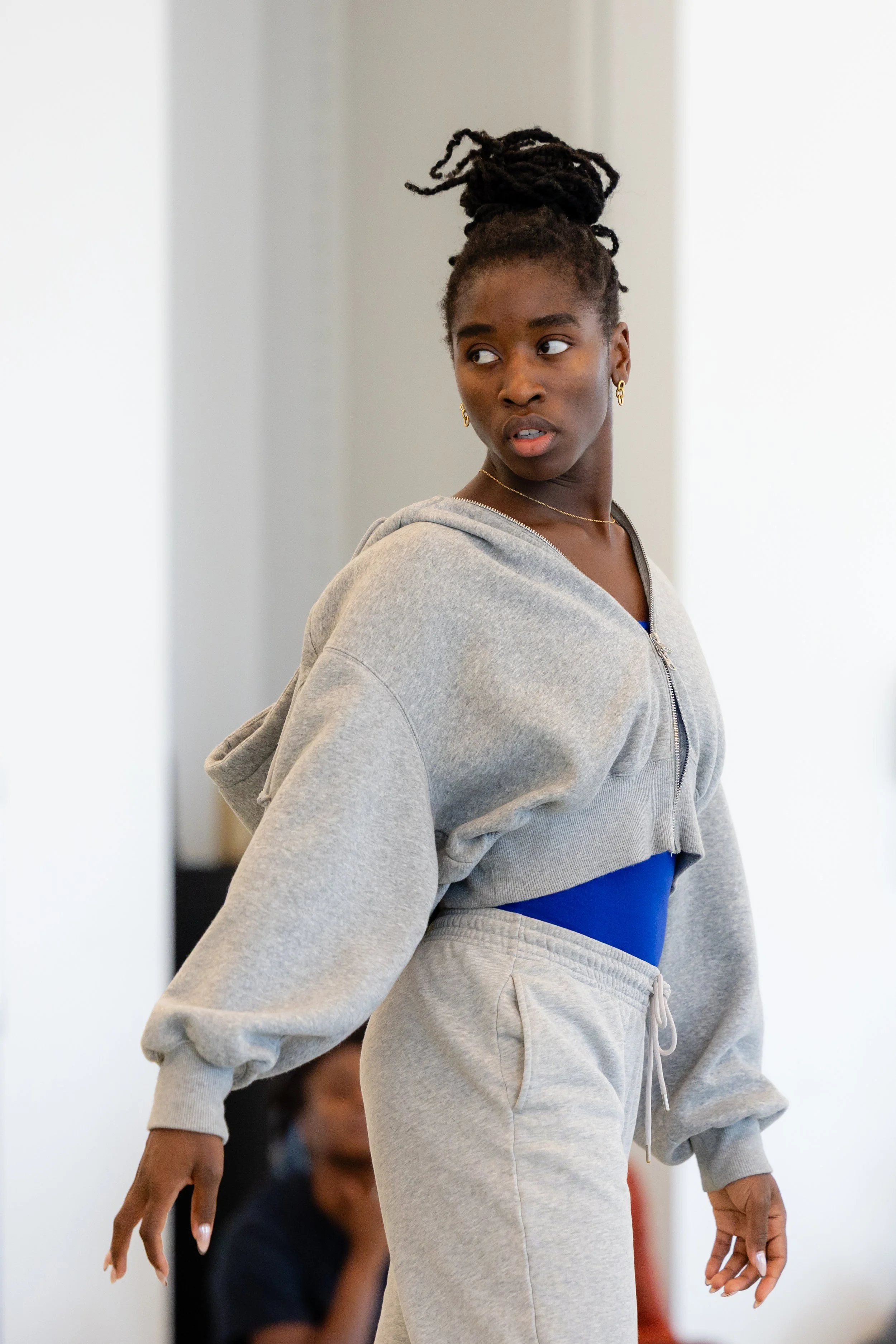 A woman with dark skin and dreadlocks styled in a high bun, wearing earrings, a gold necklace, a gray hoodie, and matching sweatpants, looks to the side with a neutral expression, in a well-lit indoor setting.