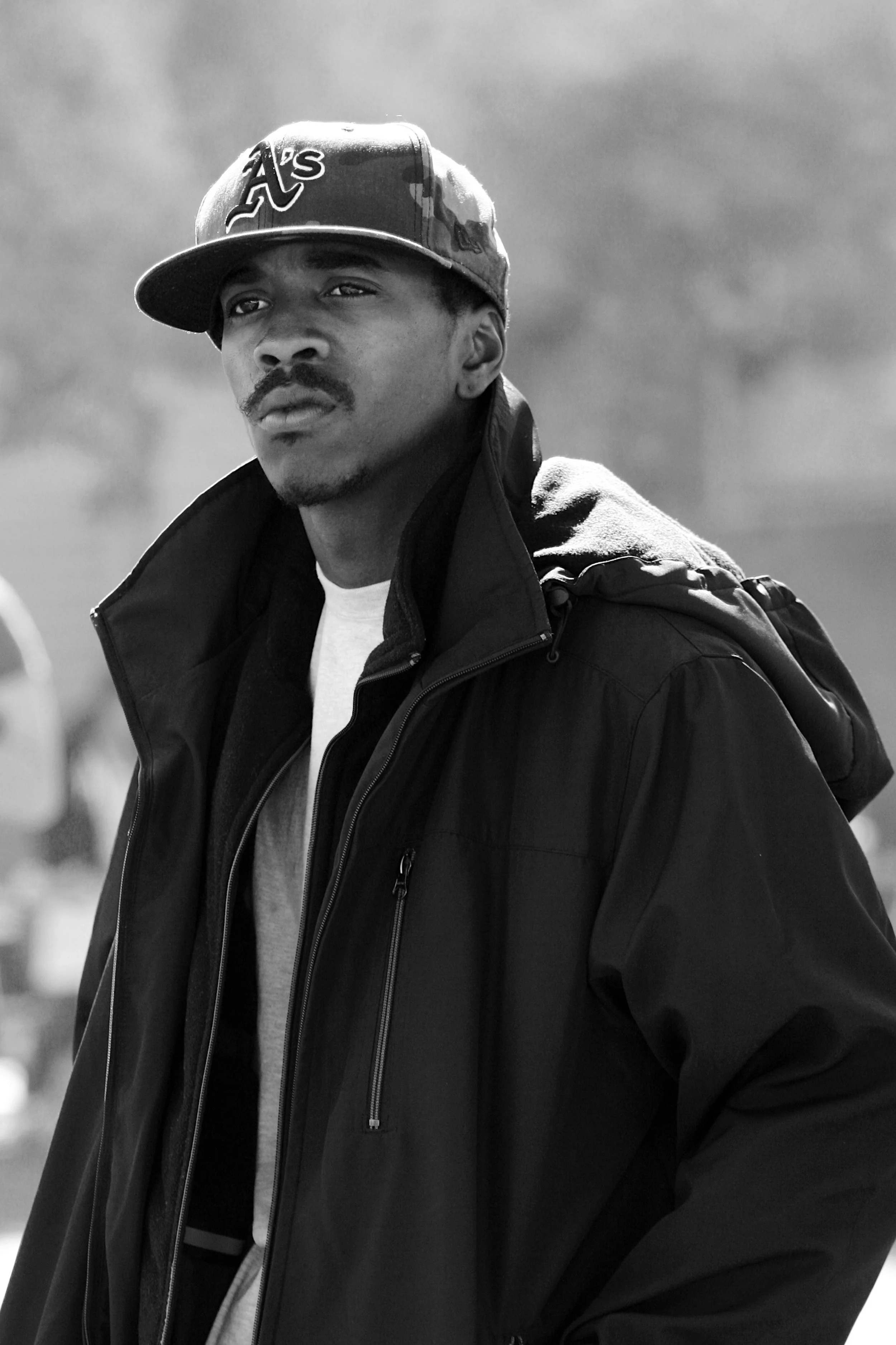 A black and white photo of a man wearing a cap with the Atlanta Braves logo, a jacket, and a white shirt, outdoors on a cloudy day.