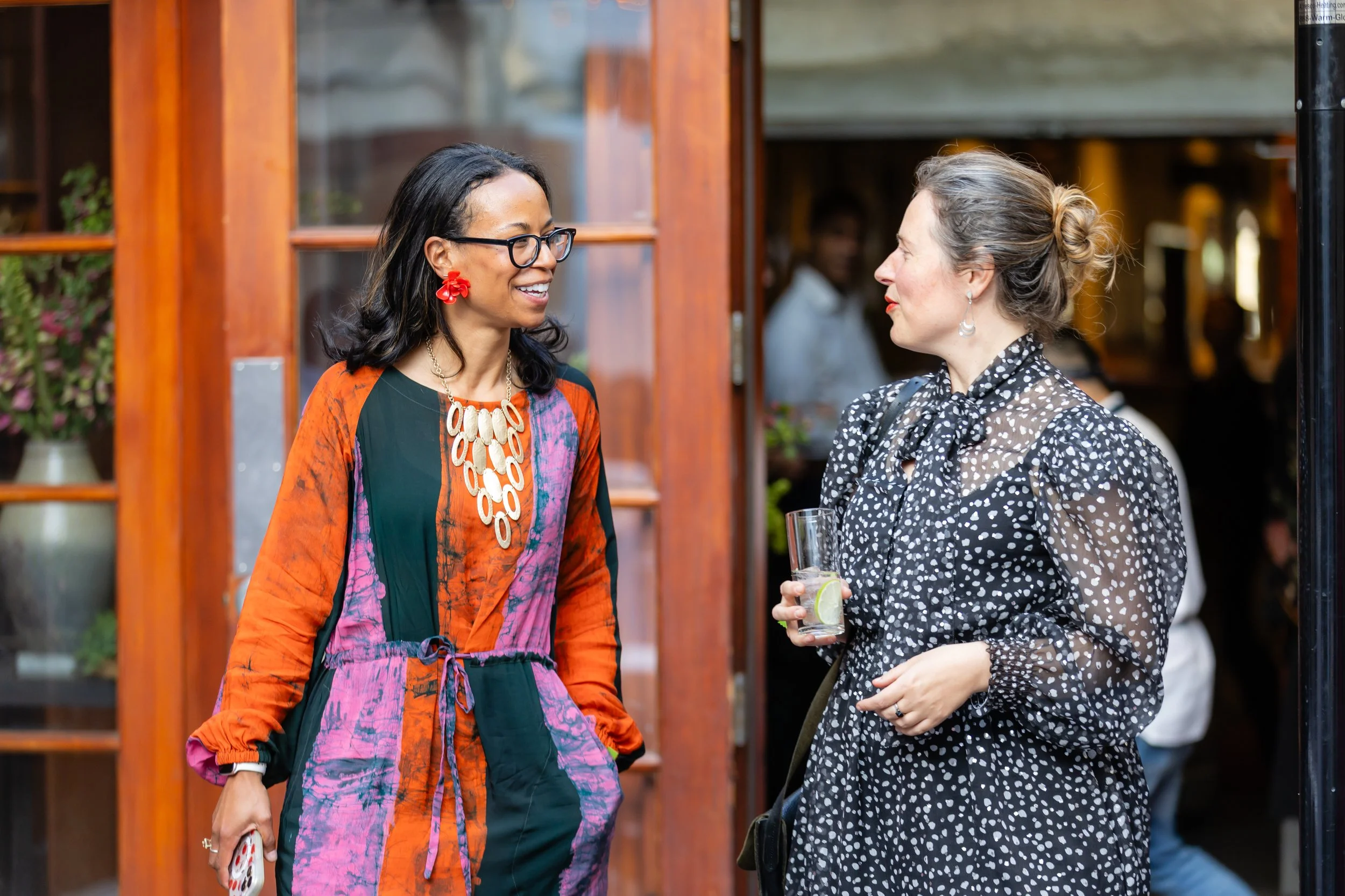 Two women are smiling and talking outdoors near a wooden structure. One woman wears glasses, a colorful dress with a bold necklace and red flower earrings, and has black hair. The other woman has blonde hair in a bun, wears a black and white polka do