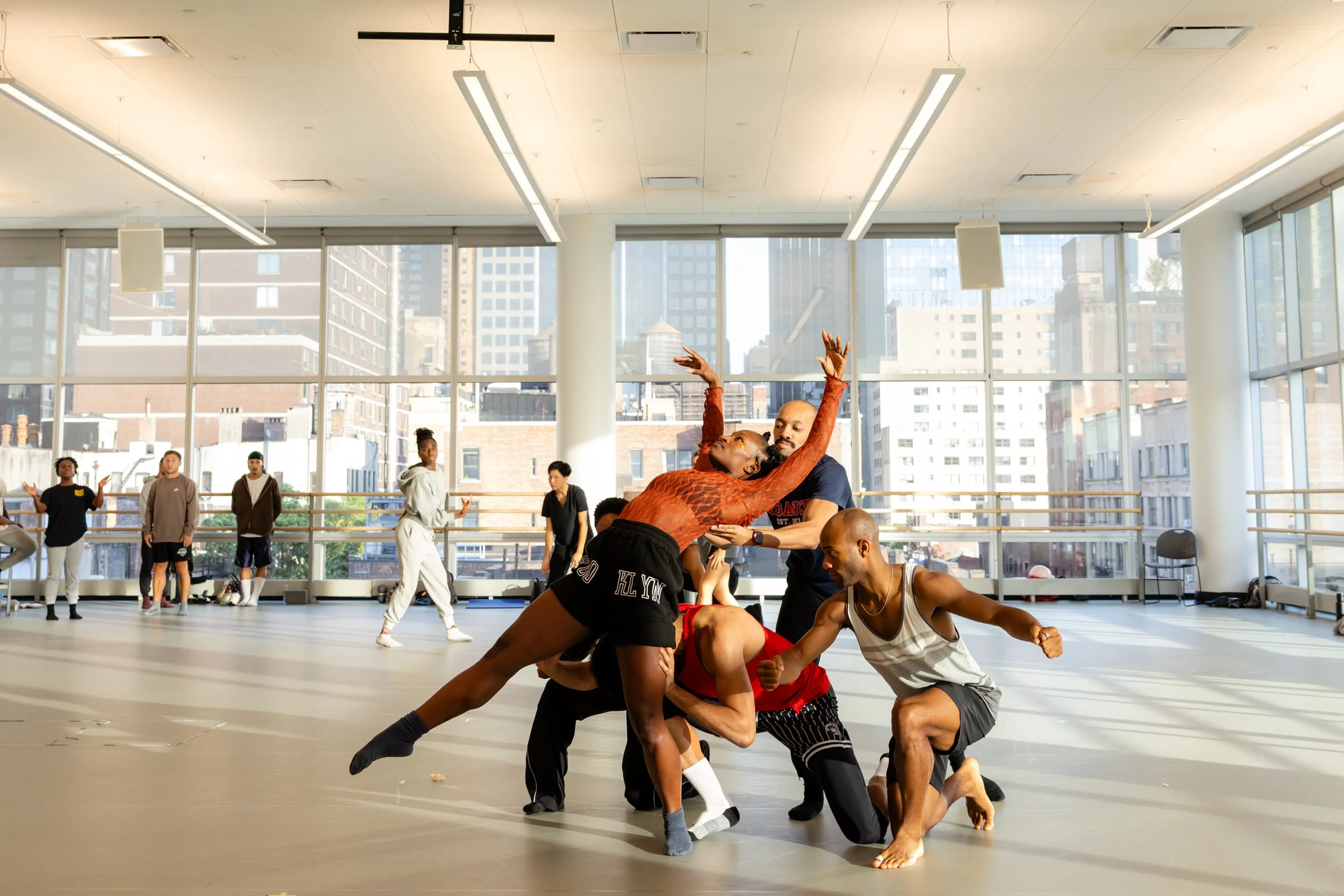 A group of dancers practicing ballet moves in a bright, airy studio with large windows revealing a cityscape. The dancers are engaged in a performance practice, with some standing and others in various poses.
