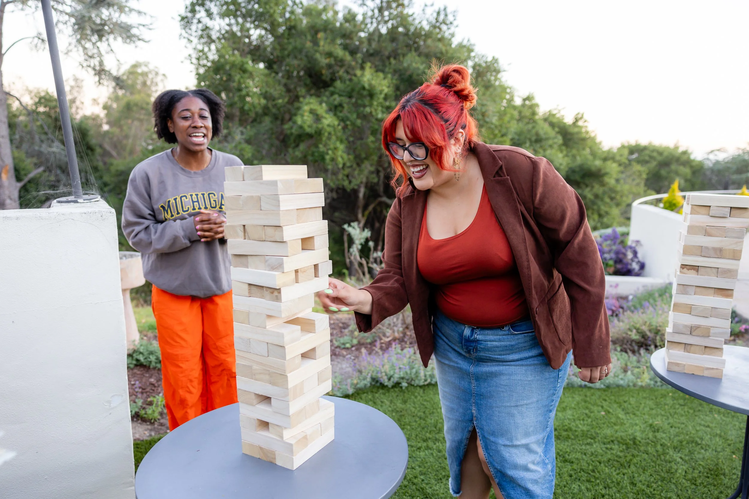Two women playing giant outdoor Jenga game with a tower of large wooden blocks on a small round table, enjoying a sunny day in a garden or park setting.