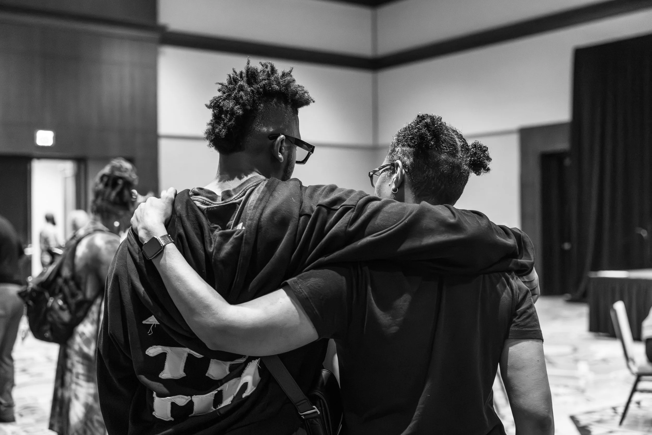 Two people hugging each other indoors, with their faces turned away from the camera. They are wearing glasses, and the scene appears to be in a public or community event setting.